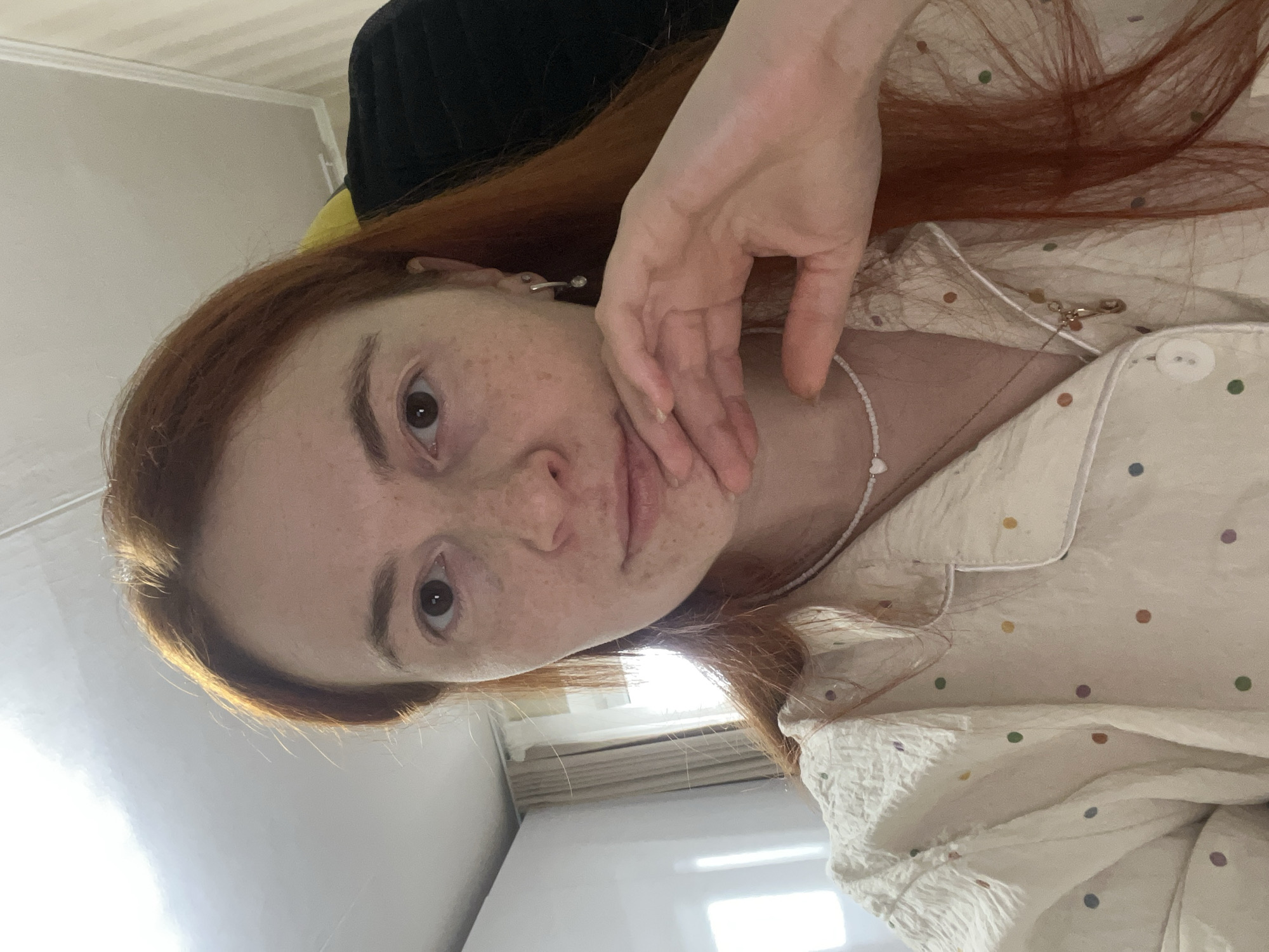A woman with auburn hair is facing the camera. Her hair partially covers the right side of her face and upper chest. She has a fair complexion with freckles. Her right hand is held against her cheek. She is wearing a light-colored top with small, colorful polka dots and a small, white beaded necklace with a heart pendant. The background includes a light-colored wall and a window with blinds, which are partially obscuring the view outside. The room appears to be indoors, with a black garment visible behind her head. The location is unknown.