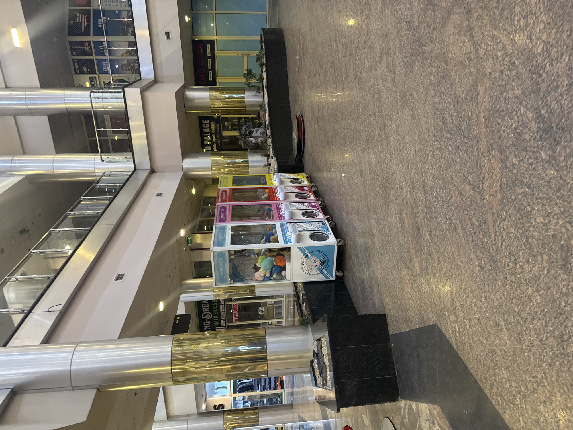 The image depicts the interior of a multi-level commercial building, likely a shopping mall, in Uyo, Nigeria. On the ground level, a row of four "Monster" branded claw crane arcade machines stands near a decorative planter area. The mall features polished stone flooring and metal support columns, with multiple storefronts visible in the background, including one for "ING-DREAM WIRELESS". Upper levels of the mall feature glass railings and additional commercial signage, including a promotional graphic displaying the flags of Australia, the UK, and the USA, along with the words "STUDY ABROAD". The mall appears to be well-lit.