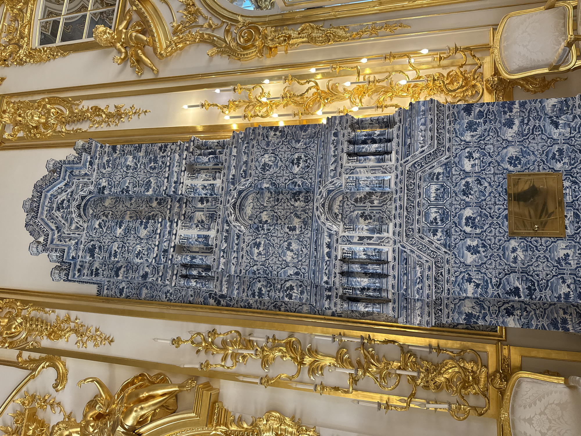 The image shows a close-up of a tall, ornate blue and white tiled stove in a richly decorated room. The stove is intricately patterned with scenes that appear to be landscapes with buildings and figures. Flanking the stove are elaborate gold-gilded wall sconces with lit candles and ornate gold-leaf decorations featuring cherubic figures and floral motifs. Two white upholstered chairs with gold frames are visible at the bottom of the image. The room's walls are white, further accentuating the vibrant blue and gold elements. This scene is likely from the Catherine Palace in Pushkin, Russia, a location renowned for its opulent interiors and decorative arts.
