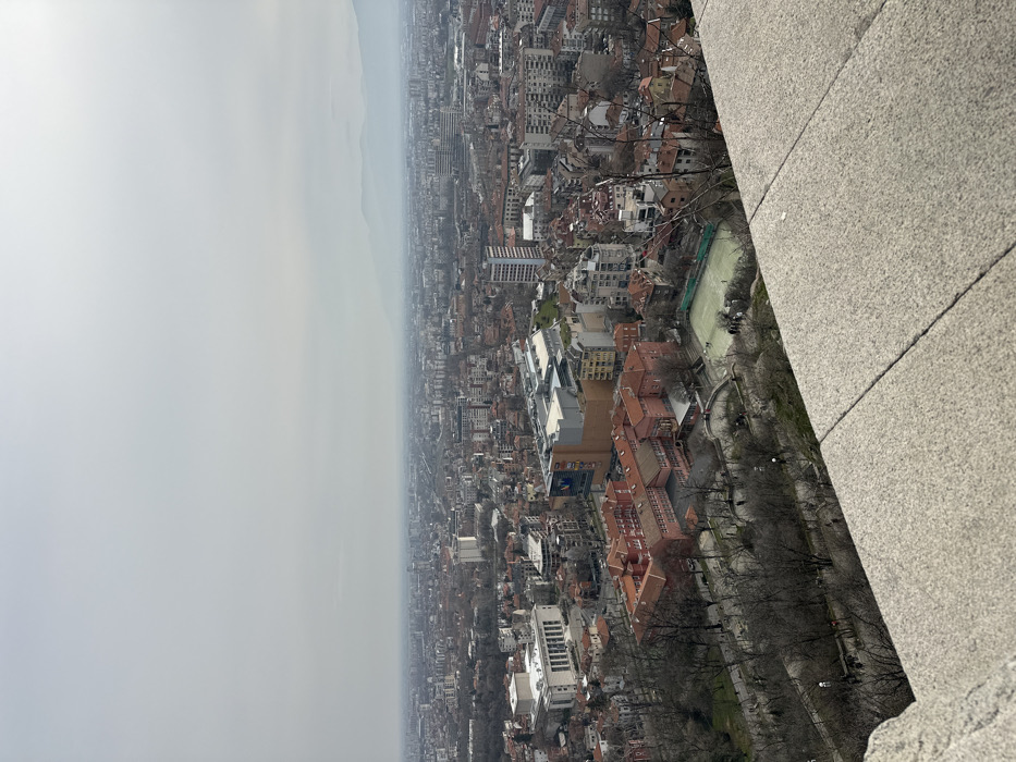The image displays an elevated panoramic view of the city of Plovdiv, Bulgaria, under an overcast and hazy sky. In the immediate foreground, the top surface of a textured, light grey stone barrier or ledge occupies the bottom right, indicating the vantage point.
Below this ledge, a steep, rocky hillside covered with bare deciduous trees descends towards the urban area. Winding pathways with visible streetlights are interspersed within this wooded slope.
In the midground, a prominent complex of buildings with red-tiled roofs and red-orange facades is visible, likely an institutional structure. Adjacent to this complex, a light-green sports court, possibly for tennis or mini-football, is discernible, enclosed by a perimeter fence. Approximately 5-7 individuals are visible on or near this court. To the left, a large, light-colored building with classical architectural elements, including columns, stands out. A modern building with a partially visible advertising banner is located further to the right.
The city extends densely into the background, characterized by a mix of residential and commercial buildings with varied heights and roof colors, primarily red-orange and grey. Several taller, contemporary buildings punctuate the urban landscape. Distant mountain ranges, partially obscured by atmospheric haze, form the horizon. The sky above is uniformly light grey and overcast. No specific actions or detailed interactions are discernible among the individuals present.