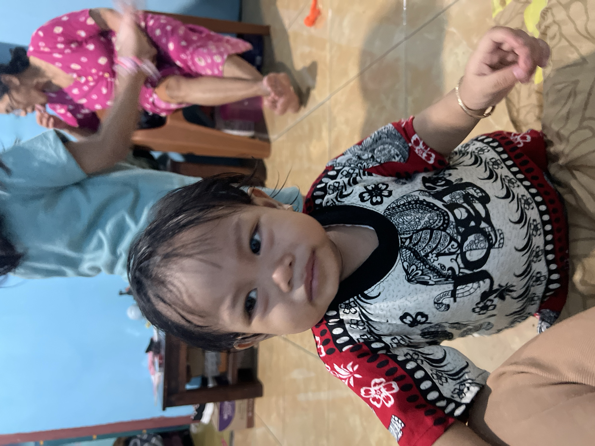 A young child with dark hair and brown eyes is the primary subject in this media file, positioned centrally. They are wearing a colorful shirt with a black collar and intricate black and white patterns, accented with red and white floral designs. To the left and slightly out of focus, an adult, presumably a woman wearing a pink top with white polka dots, is visible. The setting appears to be indoors, with a tiled floor and a light blue wall in the background. There are hints of furniture, including what looks like a wooden chair or shelf. The child's expression is neutral, looking towards the camera. The lighting suggests it is daytime. The crops highlight the child's face, their clothing, and a glimpse of the adult's attire. The overall scene depicts a casual, domestic moment.