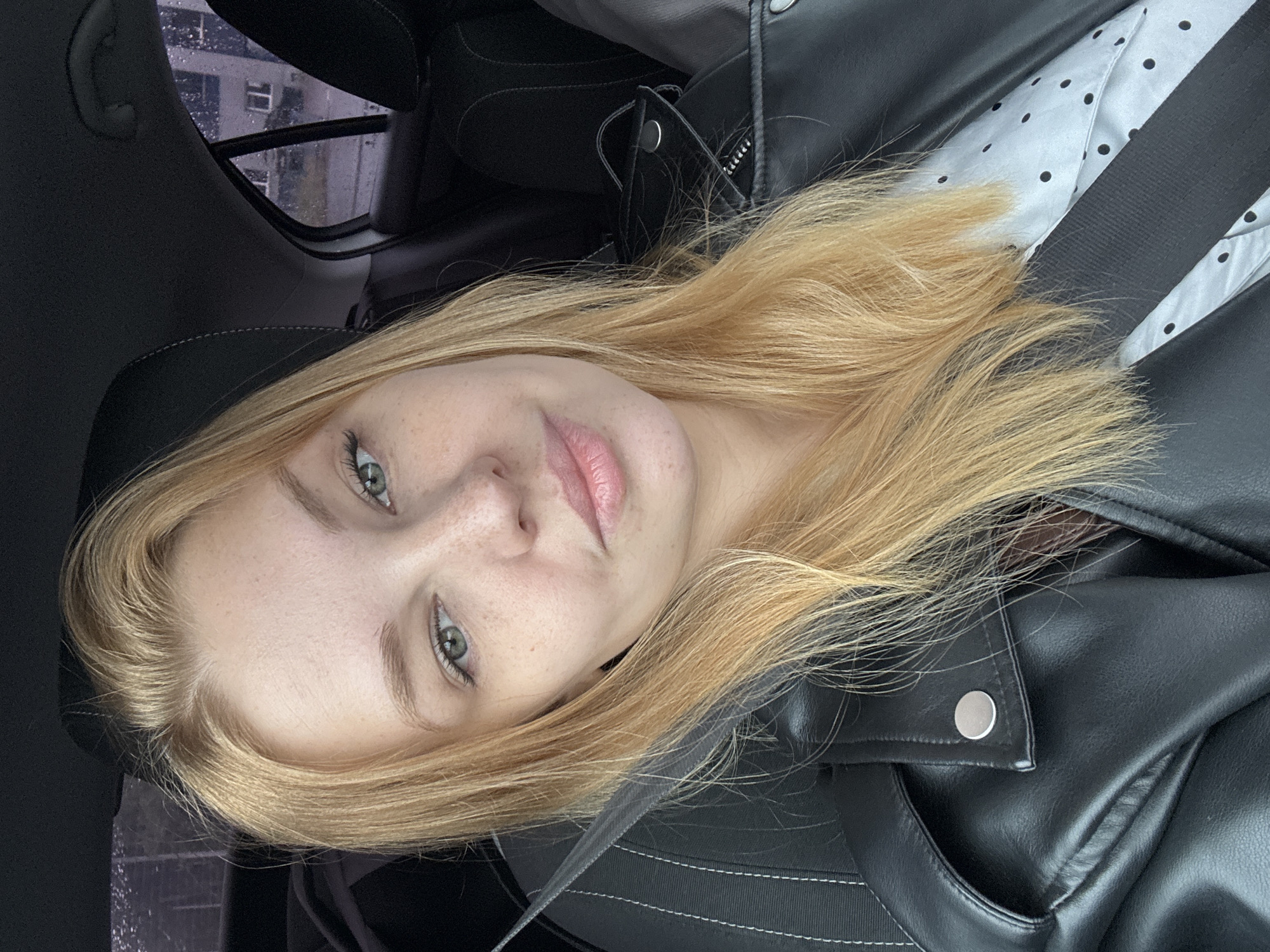 A young woman with blonde hair and light green eyes is sitting in the passenger seat of a car, wearing a black leather jacket over a white polka-dot shirt. The seatbelt is across her chest. It appears to be raining outside, with raindrops visible on the car window, through which a building can be seen. The interior of the car is dark, with black leather seats and headrests. The image is a close-up selfie, focusing on the woman's face and upper body.