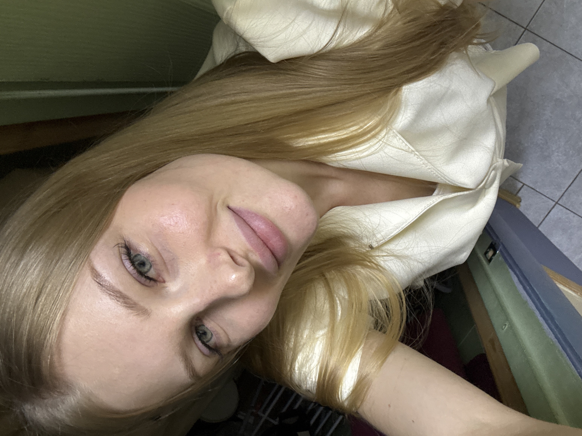 The image is a close-up, high-angle selfie of a young blonde woman with light skin and green eyes. She is wearing a light yellow or cream-colored collared shirt. Her hair is long and blonde, and falls around her face and shoulders. The background is dimly lit and appears to be indoors, with hints of furniture or shelving visible. There are no discernible landmarks or elements that would indicate the specific location within the city of Perm, Russia. The woman's face is the primary subject, with a slight smile and her gaze directed towards the camera.