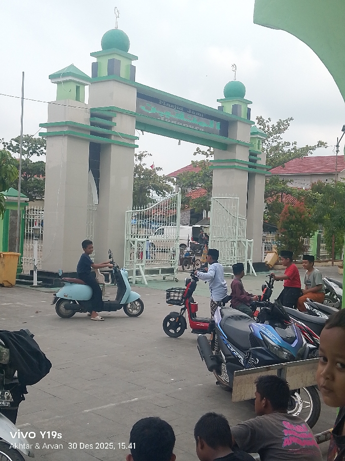 The image displays an outdoor scene in front of the Masjid Jami Jagapura mosque in Jagapura Kidul, Indonesia, under an overcast sky. A large, light-grey archway with green trim and four pillars marks the mosque entrance; each pillar is topped with a green dome featuring a crescent moon and star symbol. White metal gates are visible behind the archway, leading into the mosque grounds.
In the foreground, three boys are seated on the paved ground, two seen from behind and one's face partially visible on the right. In the midground, a group of approximately six to eight boys and young men, many wearing dark peci skullcaps, are gathered around various scooters and motorbikes. A boy in a dark t-shirt is seated on a light blue electric scooter, oriented towards the right. Another boy in a light blue patterned shirt stands near a red electric scooter, which has a front basket. Two boys are seated on a dark blue and black motorbike with a wooden platform affixed to its rear. A third boy in a maroon shirt is seated on another dark motorbike to their left. A yellow trash bin is positioned to the left of the mosque gate. In the background, houses with red-tiled roofs and trees are visible beyond the mosque entrance. The image bears a timestamp of "30 Des 2025, 15:41".
