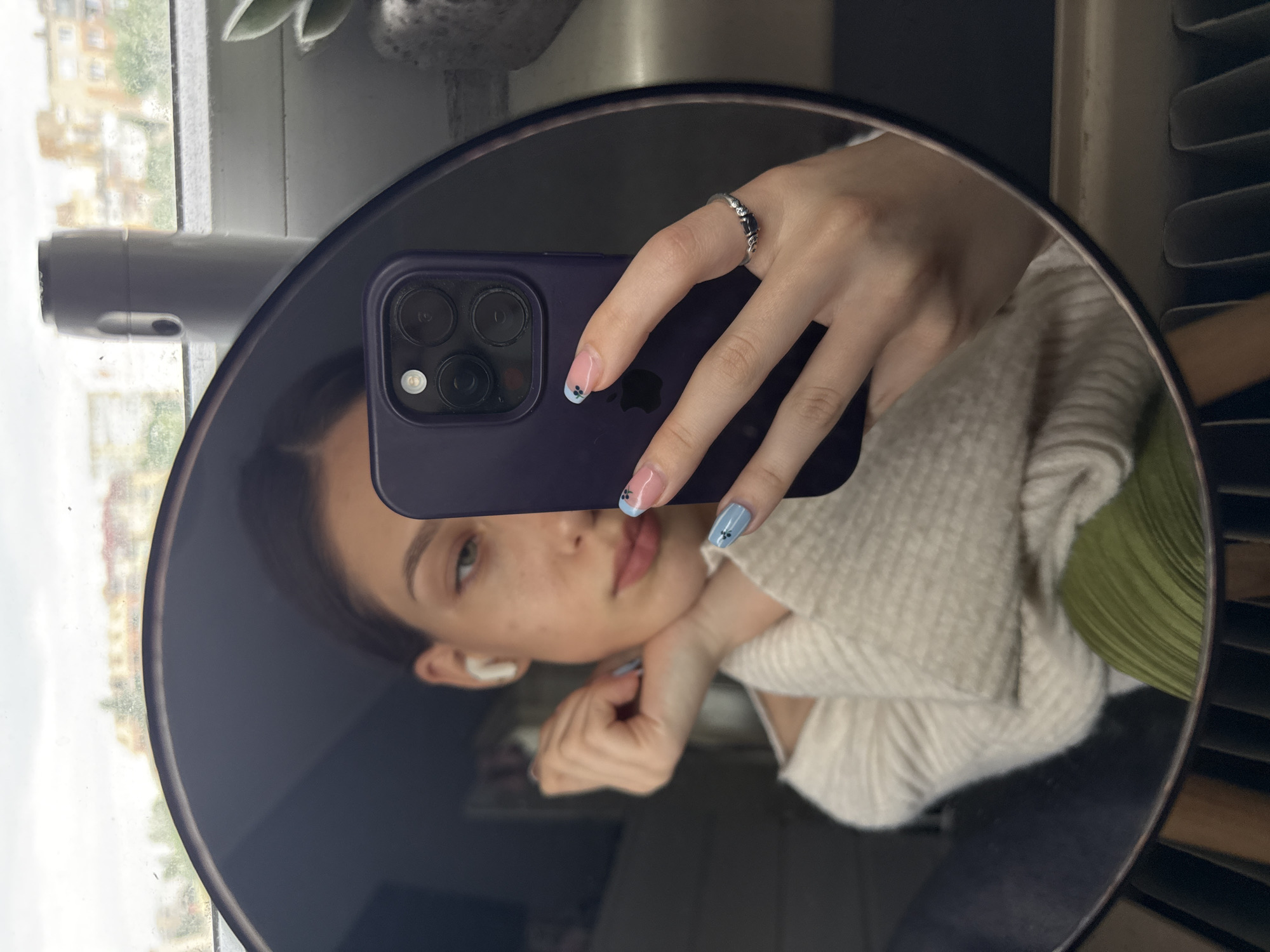 A woman is taking a mirror selfie in a round mirror. The mirror reflects her face, part of her upper body, and her hands holding a dark purple smartphone. She is wearing a light-colored sweater and has an earbud in one ear. Her nails are painted with a light blue and pink design. She is wearing a silver ring. The reflection also shows a window with a view of buildings and a plant, as well as a radiator. The exact location in Sofia, Bulgaria cannot be determined from this image.