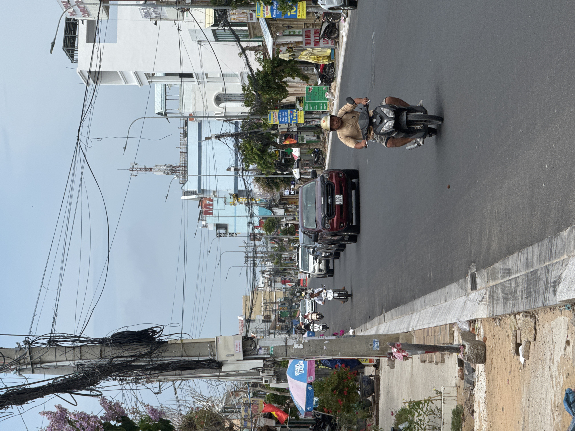The image depicts a street scene in Phan Thiết, Vietnam, characterized by a relatively narrow asphalt road lined with buildings and dense overhead utility lines. In the foreground, a man wearing a light brown t-shirt and shorts rides a black motorcycle, heading towards the viewer. His face is partially obscured by a helmet. The road is moderately busy with several other motorcycles and cars. To the left of the road, a sidewalk area is visible with a utility pole heavily laden with black wires, a pink and blue umbrella, and some potted plants. Construction debris and sand are also present on the left side. The buildings on the right are multi-story, with a mix of modern white structures and older facades. A cell tower can be seen in the distance. The sky is overcast. The overall impression is one of a bustling, somewhat chaotic urban environment typical of a Vietnamese city.