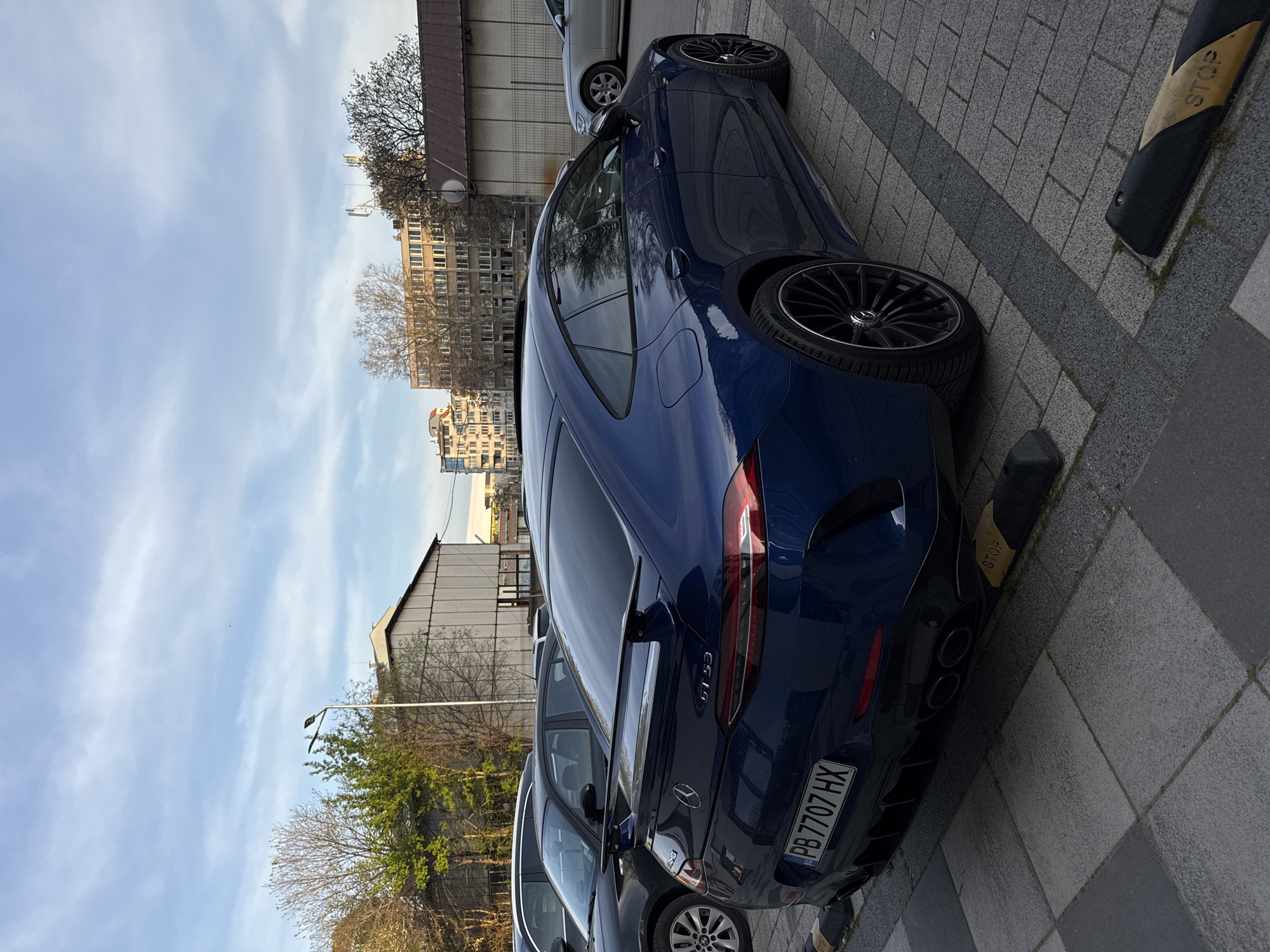 The image shows a navy blue Mercedes-AMG GT 53 4-Door Coupé parked on a paved surface. A license plate is visible, reading "PB 7707 HX". The car is parked next to a street border marked with "STOP". In the background, there are buildings, trees and parked cars. Based on the license plate, the car could be registered in Plovdiv, Bulgaria.