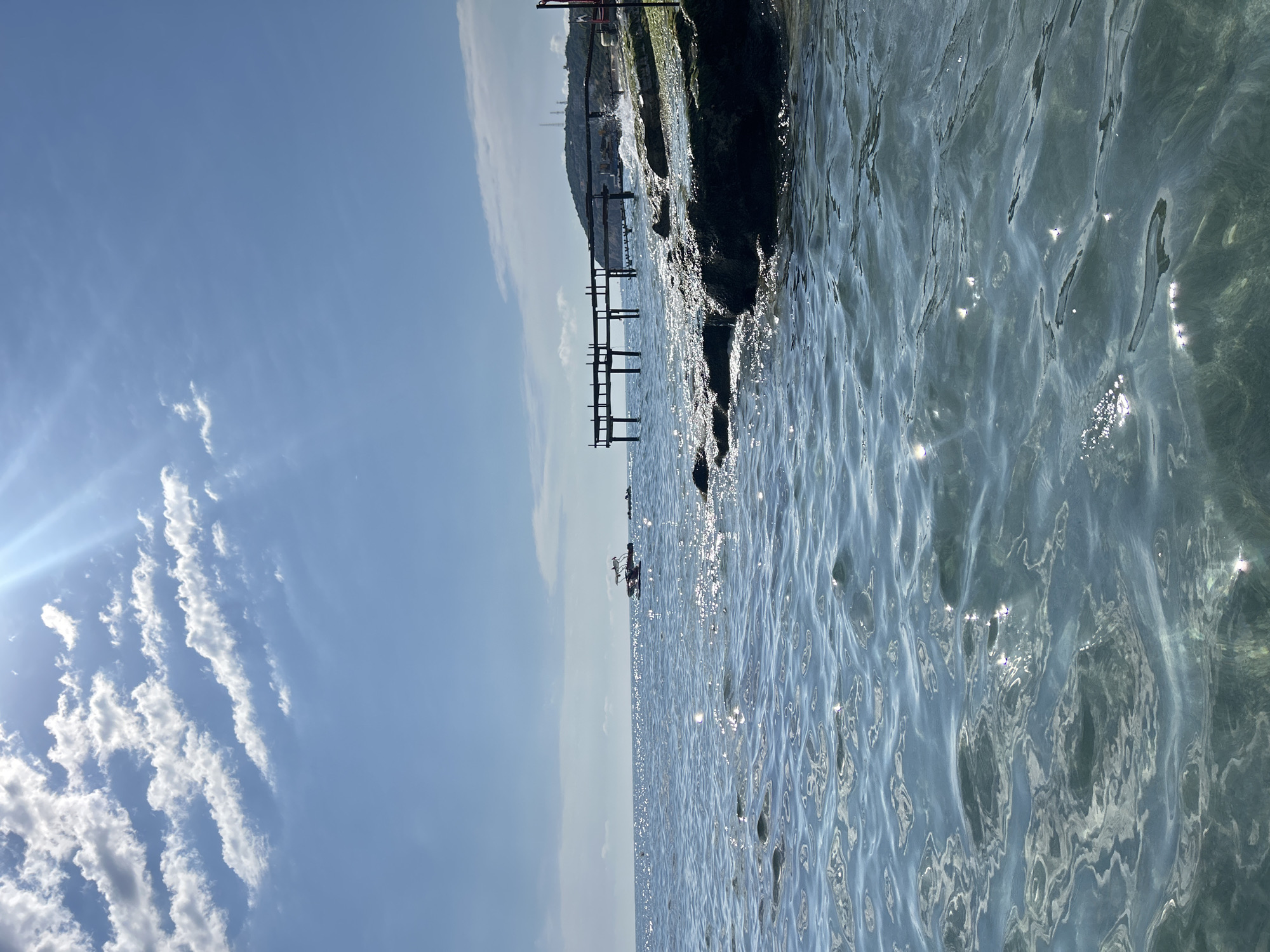 The image depicts a body of water, likely a sea or large lake, under a partly cloudy sky. The water surface is shimmering with sunlight, indicating clear conditions. A small boat or watercraft with one or two individuals is visible in the middle distance. A wooden pier or structure extends into the water from a rocky shoreline in the background. The location is likely near a coastline or lakeside. There is no data on the city.