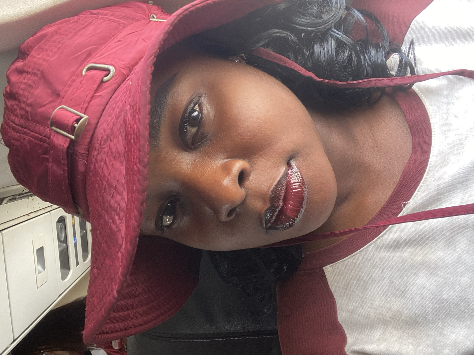 A close-up portrait features a Black woman captured from an angled perspective. She has dark skin and dark, curly hair visible around her face. She wears a maroon or dark red bucket hat with metallic buckles and adjusters on its side, and a matching strap hangs loosely around her neck. Her lips are covered in a glossy, dark red lipstick. She is dressed in a t-shirt that has a white body and maroon trim around the collar, coordinating with her hat. Her gaze is directed confidently towards the camera.
The background is an indoor setting, showing blurred details of what appears to be a vehicle interior, possibly public transport in Ikeja, Nigeria. White panels or cabinets with handles and reflections are discernible, along with a dark surface, potentially a seat. The lighting is bright and even, highlighting the subject's features. No text is visible in the image.