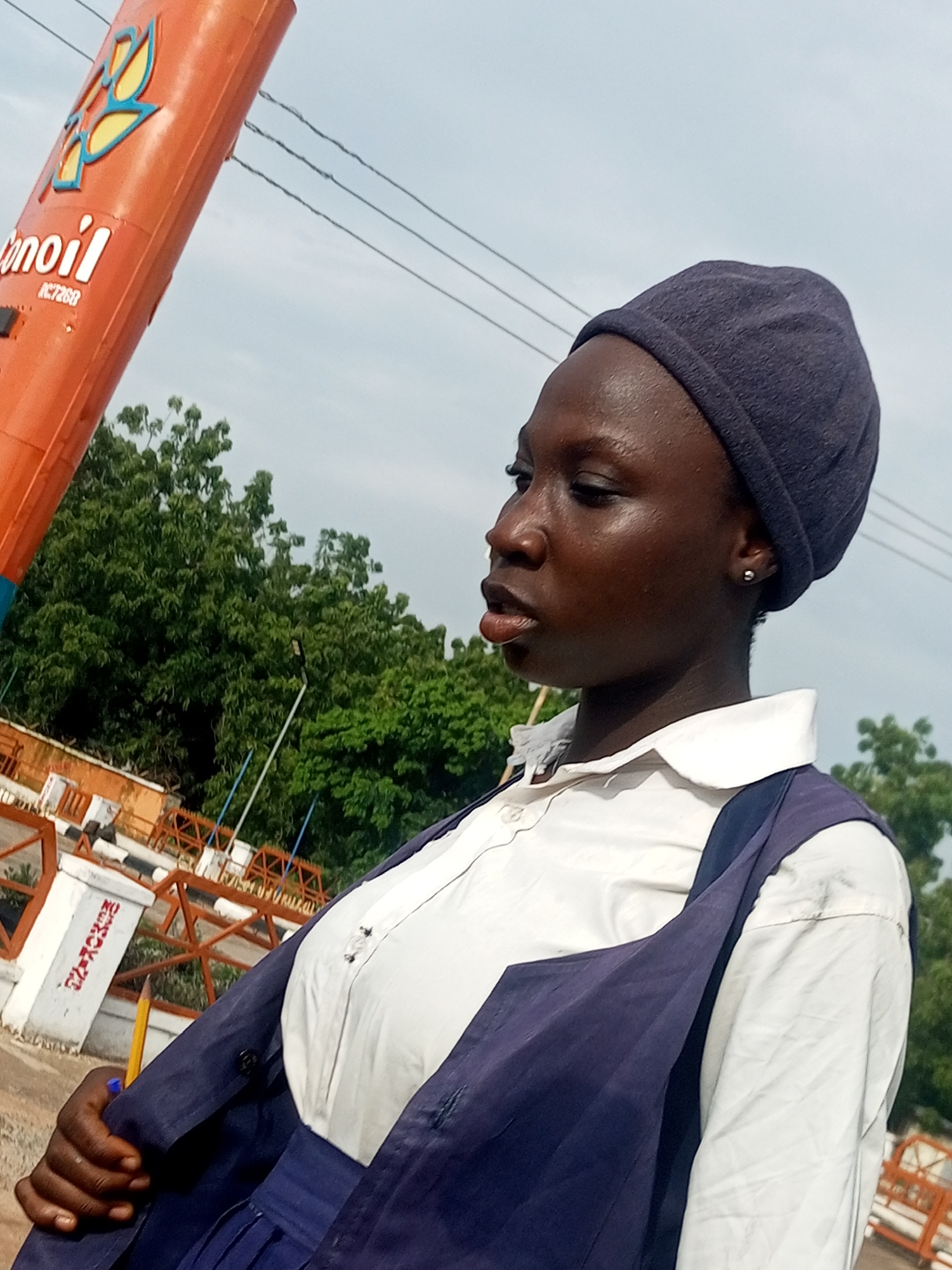 A young Black person wearing a dark blue headscarf, a white collared shirt, and a dark blue vest stands outside in Gombe, Nigeria. The person is looking to their left with their mouth slightly open. In the background, there is an orange pylon with the logo and text "conoil" and "RC7268" on it. Behind the person and the pylon, there are trees and a cloudy sky. The person is holding a yellow pencil in their right hand.