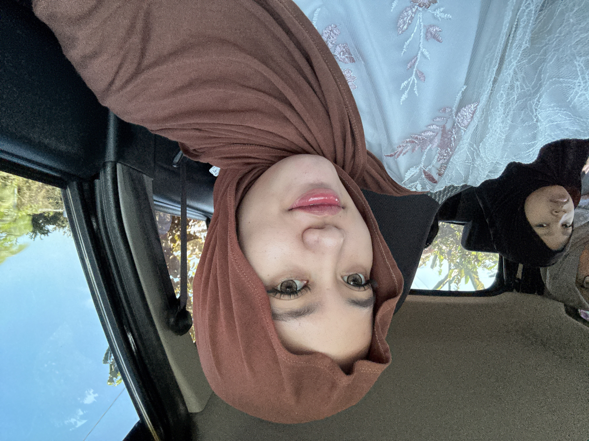 This is a selfie taken from inside a car, with three women visible. The woman in the foreground is wearing a brown hijab and a white dress with pink floral embroidery. She is looking at the camera with her eyes open and lips slightly parted, wearing makeup including eyeliner and lip gloss. To her left, in the backseat, a younger woman is visible wearing a black hijab and looking slightly to the side. The third woman, partially obscured, is wearing a grey hijab. The interior of the car is visible, including the headliner and the side door panel. Outside the car windows, lush green trees and a bright blue sky are visible, suggesting a rural or suburban setting, possibly in Gadingrejo, Indonesia, given the context. The lighting suggests it is daytime, likely sunny.