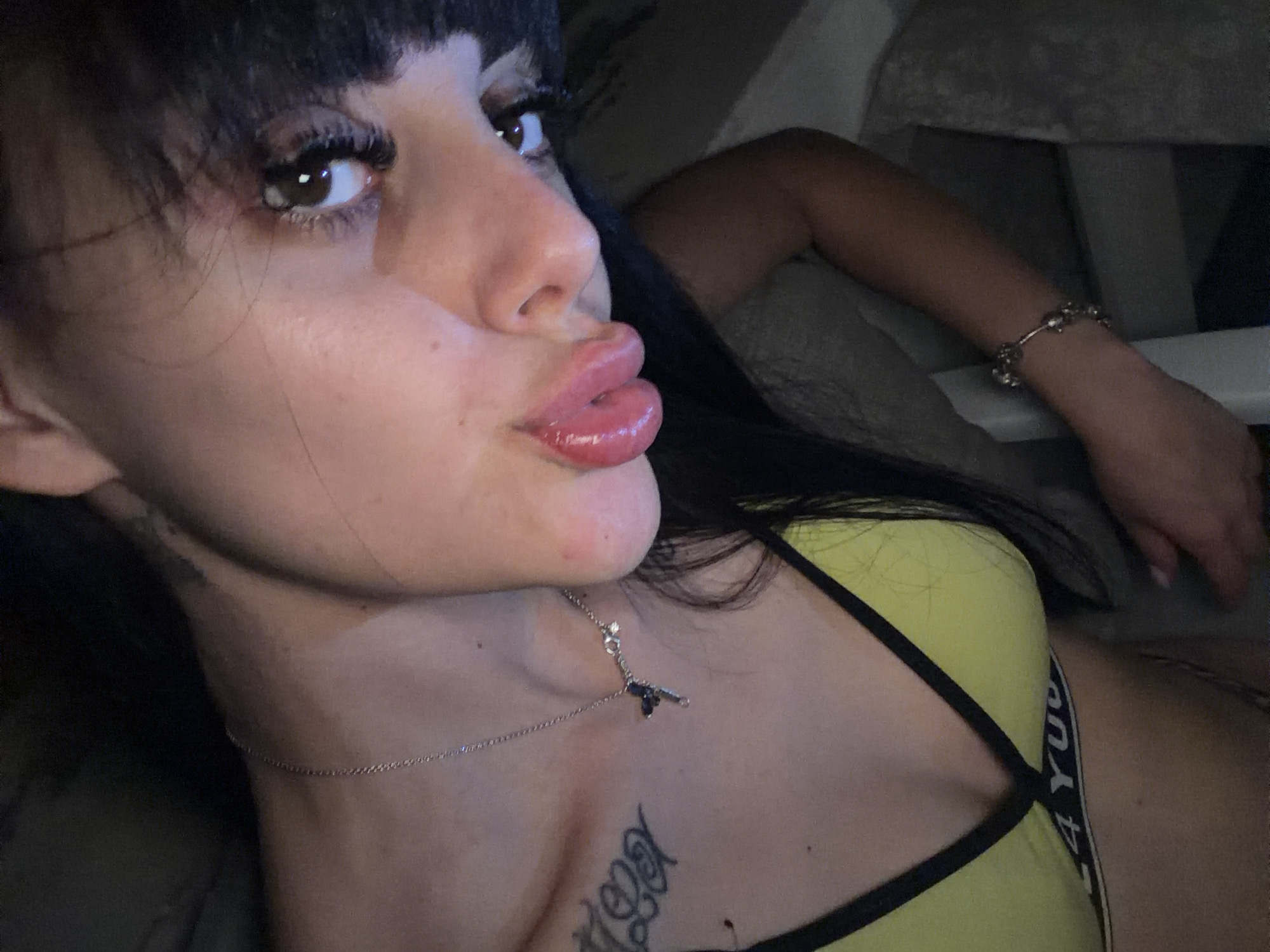 The image shows a close-up, angled shot of a young woman. She is wearing a bright yellow bikini top with black trim. A silver chain with a dark pendant hangs around her neck, resting on her chest. She has dark hair, some of which falls forward, and appears to be wearing makeup, including mascara and lip gloss. A tattoo is visible on her upper left chest. She has a bracelet on her right wrist. The background is dark and out of focus, making it difficult to discern the environment. There are no discernible features that place the image in Plovdiv, Bulgaria.