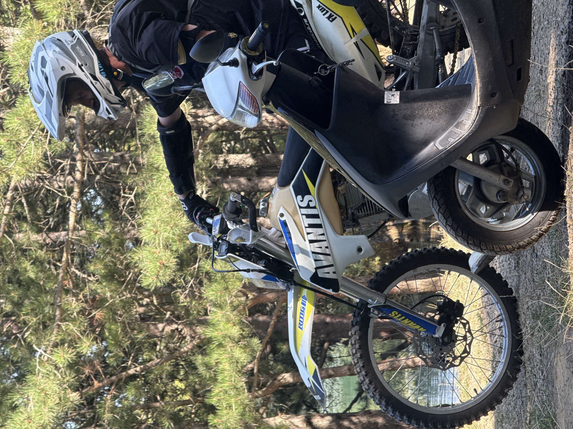 In Zaprudnoe, Ukraine, a person wearing a motocross helmet and protective gear sits on a dirt bike. The dirt bike is parked next to a scooter. The vehicles are surrounded by trees and grass, with a body of water visible in the background. The scene appears to be outdoors, possibly a recreational area or a trail.