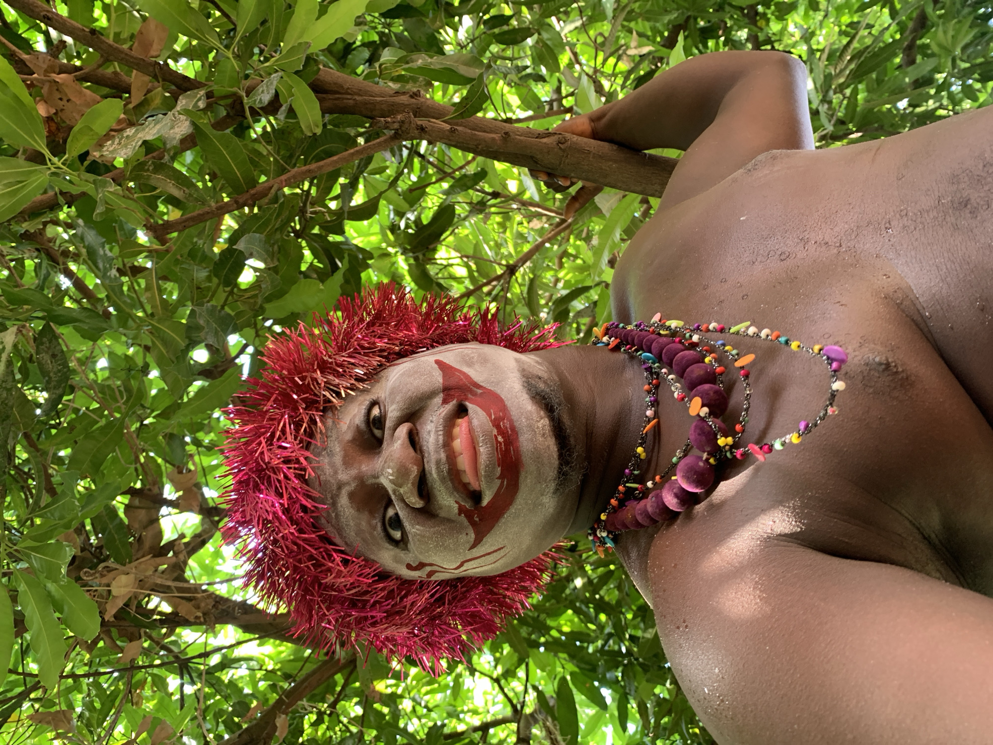 The image, taken in an outdoor, natural environment in Akwanga, Nigeria, features a dark-skinned man in traditional attire. He is partially visible from the chest up, positioned within the branches of a tree, which frame the shot with green foliage. The man wears a red, fringed headdress, and his face is painted with white makeup around the eyes and mouth, forming a clown-like design. He also sports a colorful necklace with a variety of beads, including large, round, purple ones. His upper body is bare, and he is holding onto a tree branch with his right hand. The image is shot from a low angle, looking up at the subject.