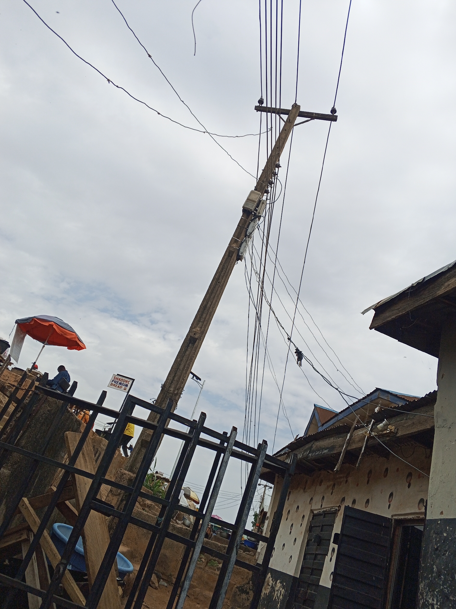 The image, taken in what is likely Gwong, Nigeria, showcases a somewhat dilapidated urban scene. A leaning wooden utility pole dominates the view, with numerous electrical wires strung haphazardly. At the top of the pole, two birds are perched. A small rectangular box is attached to the pole. The sky is overcast with gray clouds.
On the left, behind a black metal fence, is an orange and gray parasol. A person wearing a blue shirt is seated beneath it. A sign reading "SHADDRE PREMIUM BREAD" is also visible near the fence.
To the right, a partially visible building reveals deterioration. The walls are stained and marked, and the roof is damaged. A window with dark shutters is visible, as is the partially open door.