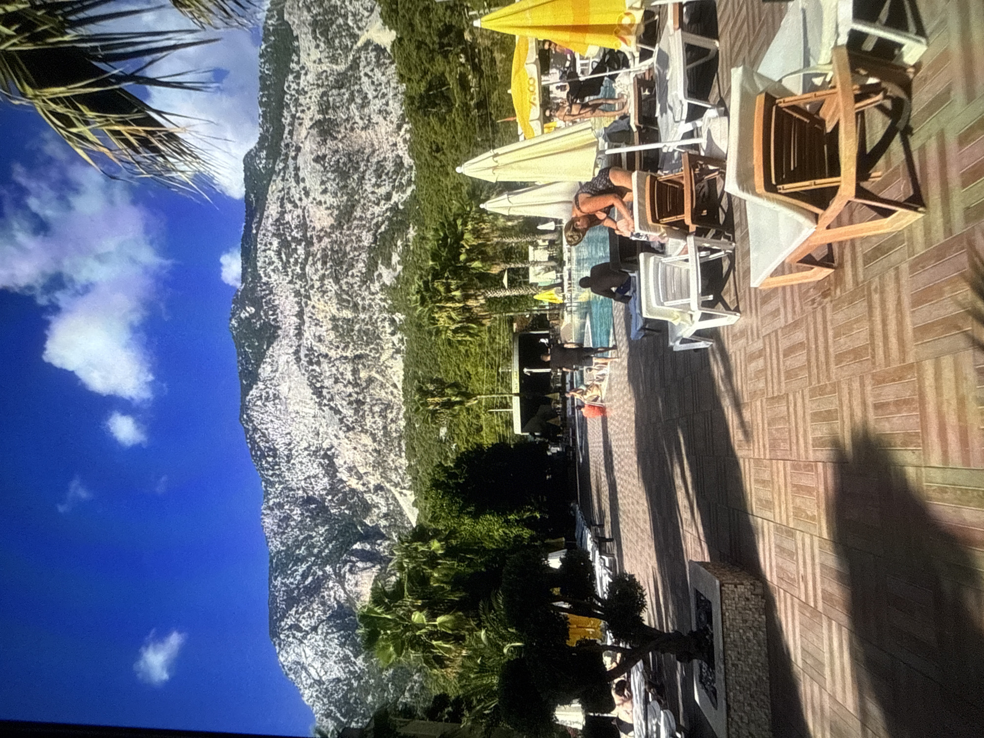 The image depicts a sun-drenched outdoor scene with a focus on a swimming pool area. A large, imposing mountain with a rocky texture dominates the left side of the frame, juxtaposed against a vibrant blue sky with scattered white clouds. Lush green foliage, including palm trees, lines the base of the mountain and extends towards the pool.
The pool area itself is paved with a patterned wooden deck. Numerous lounge chairs, some occupied by people, are arranged around the pool. Several yellow and white umbrellas provide shade. A woman in a dark swimsuit is visible tending to something on a lounge chair. Other individuals are seen relaxing on chairs or walking in the background. The scene suggests a leisurely daytime activity, likely a vacation or a relaxing day at a resort.
The time of day appears to be late afternoon, indicated by the long shadows cast across the wooden deck. The weather is clear and sunny, suitable for outdoor relaxation. While the image does not provide explicit text, the overall atmosphere is one of tranquility and summer enjoyment. The setting appears to be a resort or a hotel with a pool and a view of a mountainous landscape. The presence of lounge chairs, umbrellas, and a swimming pool strongly suggests a leisure-oriented environment.
