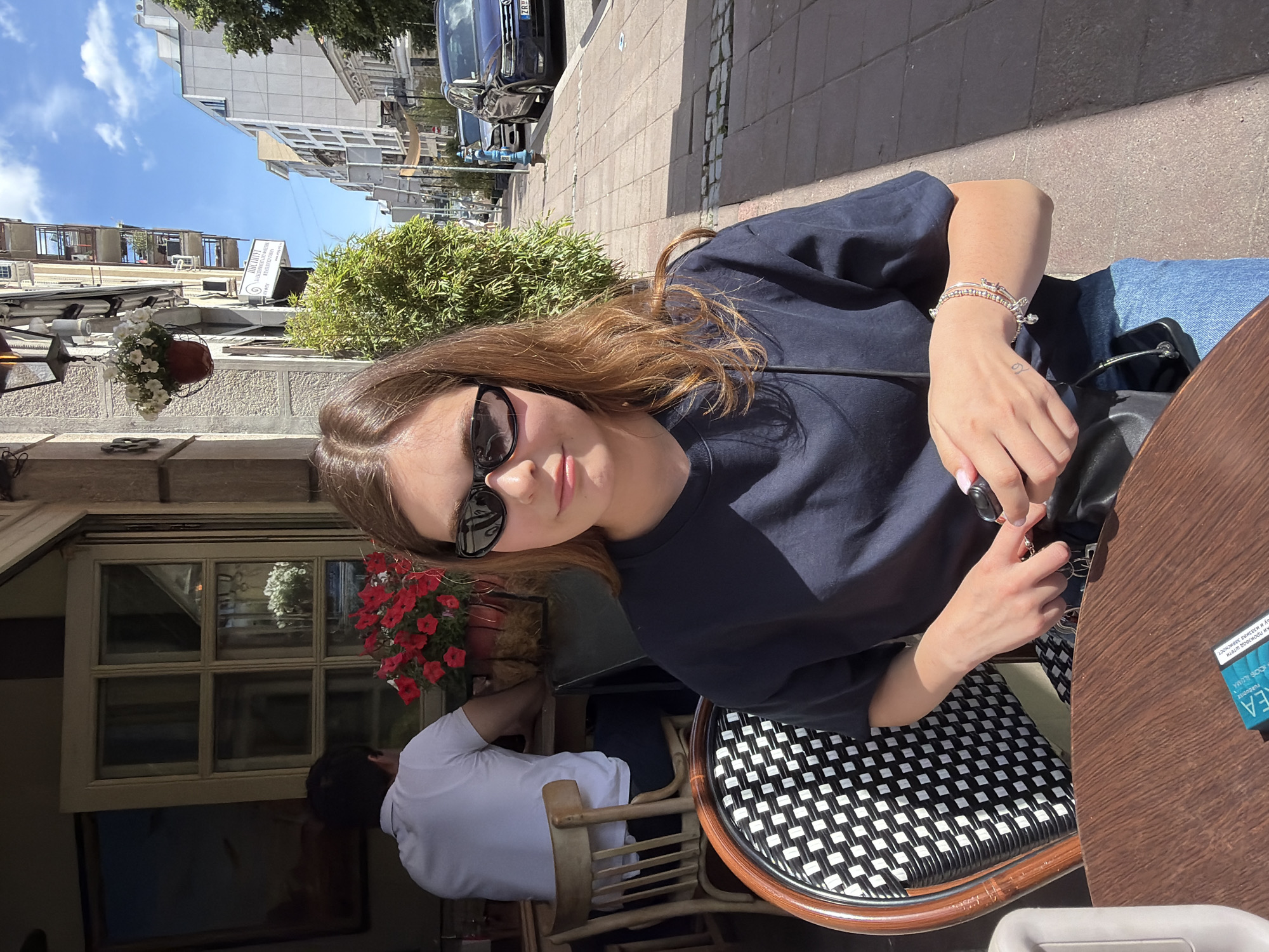 A woman wearing sunglasses and a dark blue t-shirt sits at a cafe with a black and white woven chair on a sunny day. She is seated at a wooden table, holding a black leather bag. On the table to her right, there is a blue package, potentially cigarettes. Behind her, a man in a white shirt is seated at a table inside the cafe, next to a window display with red flowers. Outside, the buildings, streets, and cars of Belgrade, Serbia are visible, suggesting an urban setting.