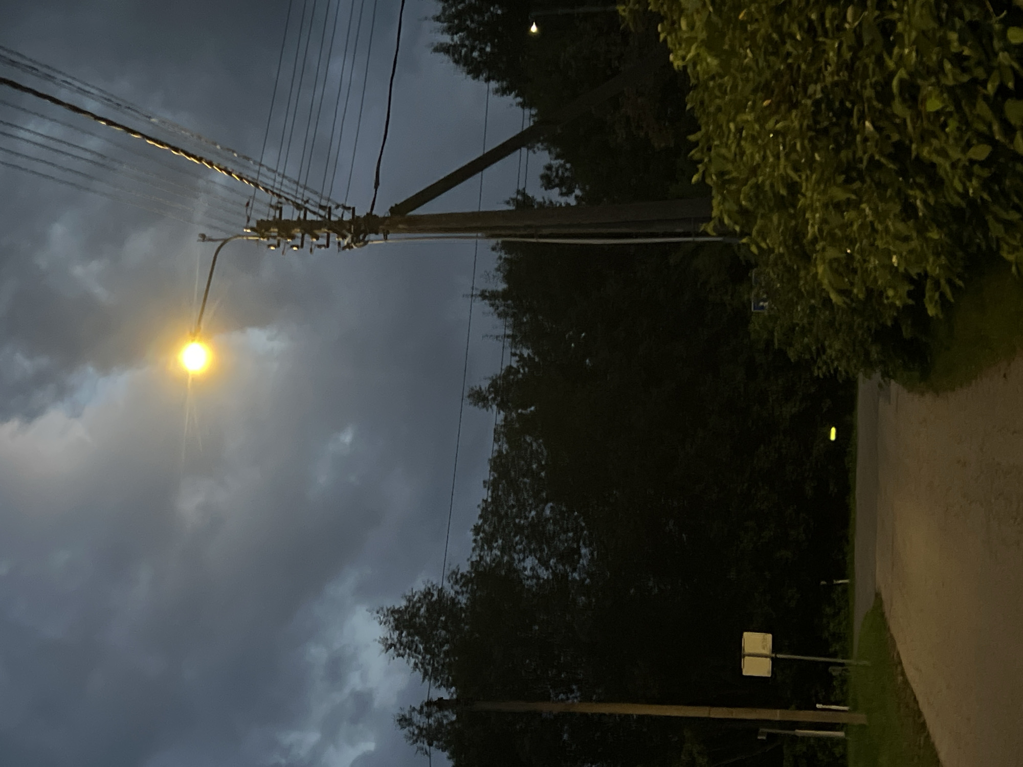 A street lamp illuminates a dimly lit, rural road on a cloudy evening. The road curves to the left and disappears into the darkness, bordered by lush green bushes and trees. A power line pole stands to the right of the road, carrying multiple wires overhead. In the distance, another street lamp casts a faint yellow glow. The sky is heavy with dark, dramatic clouds, suggesting impending rain or a recent storm. The overall atmosphere is serene and quiet, with no people or vehicles visible in the scene.