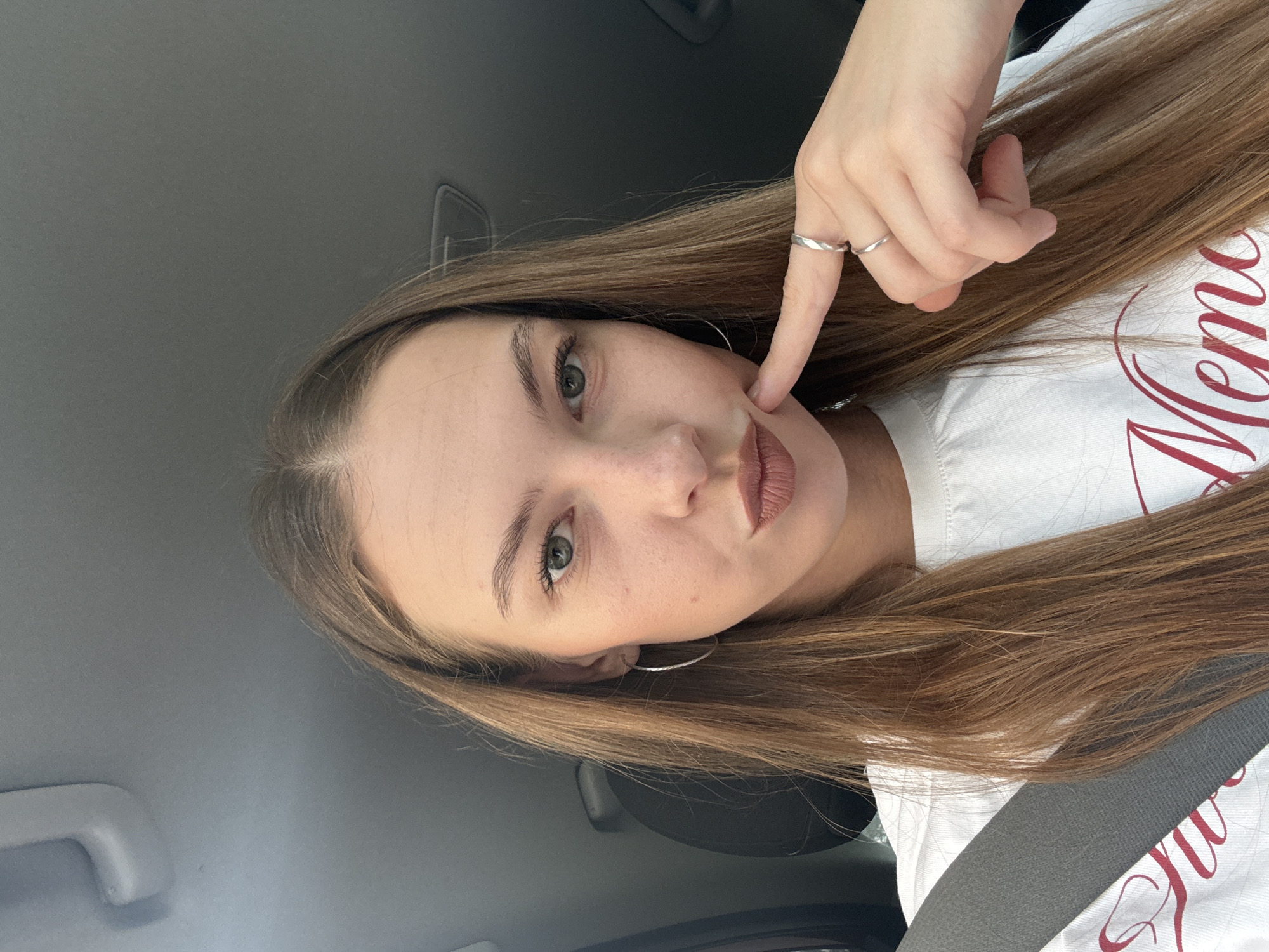 A young woman with long, straight, brown hair and green eyes is taking a selfie in a car. She is wearing a white t-shirt with red lettering and a seatbelt is visible across her chest. She has a silver hoop earring in her left ear and is wearing two silver rings on her right hand, which she is using to gently touch her cheek. The background of the image is the interior of a car. The location is not specified.