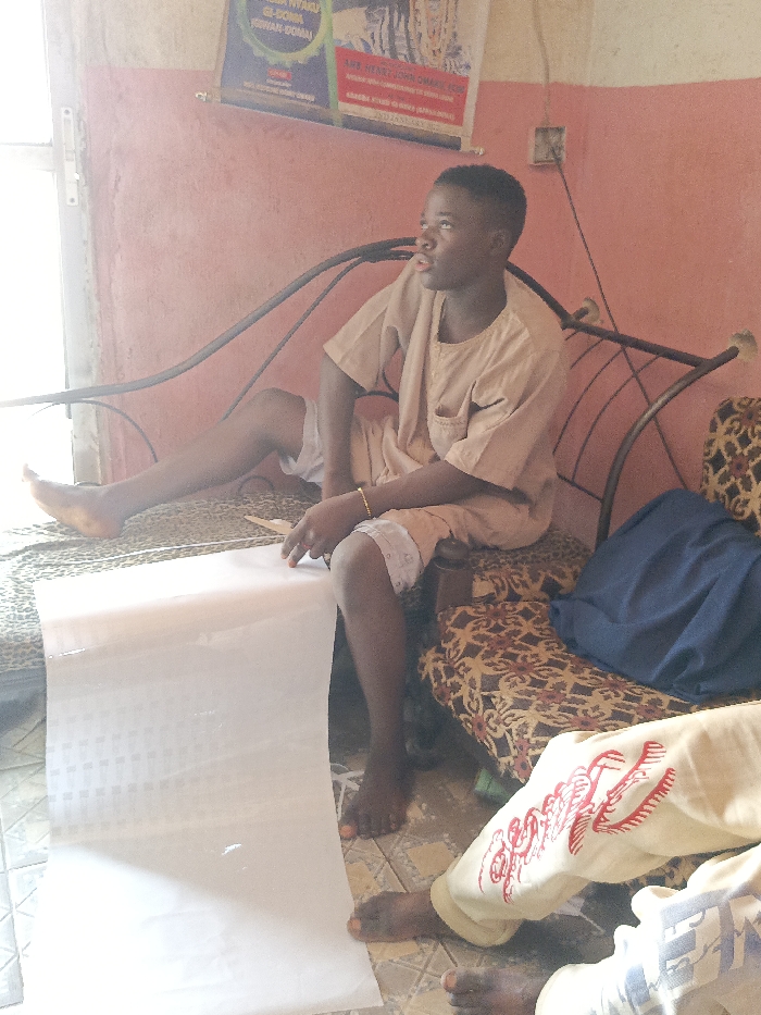 A young man sits on a metal-framed sofa, wearing a short-sleeved shirt and shorts. He holds a roll of white paper, looking to the side with a thoughtful expression. Another person, partially visible, sits beside him, wearing pants with red embroidery. The sofa has patterned cushions and a blue cloth draped over it. The background includes a faded pink wall, a partially visible doorway, and framed posters. The scene is indoors.