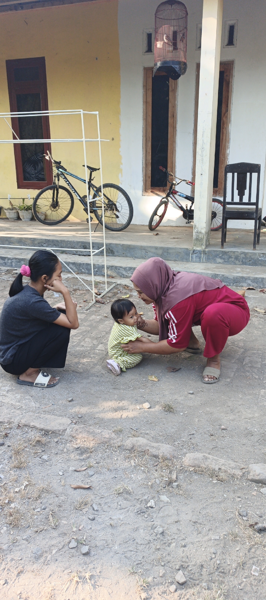 In Kasiyan, Indonesia, three individuals are positioned in an outdoor setting with a building in the background. A young girl, wearing a grey t-shirt and black pants, is crouched on the ground, her head tilted downwards. Beside her, an adult woman, draped in a maroon hijab and a matching maroon outfit with white stripes on the sleeves, is also crouched, interacting with a baby. The baby, dressed in a green and white striped outfit and light pink shoes, is sitting on the ground and facing the woman. The environment consists of a dirt ground with scattered leaves and small stones. In the background, a yellow-walled building with wooden doors and windows is visible. A white metal rack stands to the left of the building, with a black and blue bicycle leaning against it. Further to the right, another bicycle, primarily red, is propped up. A dark wooden chair is placed near the building's entrance. A birdcage, containing at least one bird, is suspended from a pole. The overall scene suggests a casual outdoor gathering.