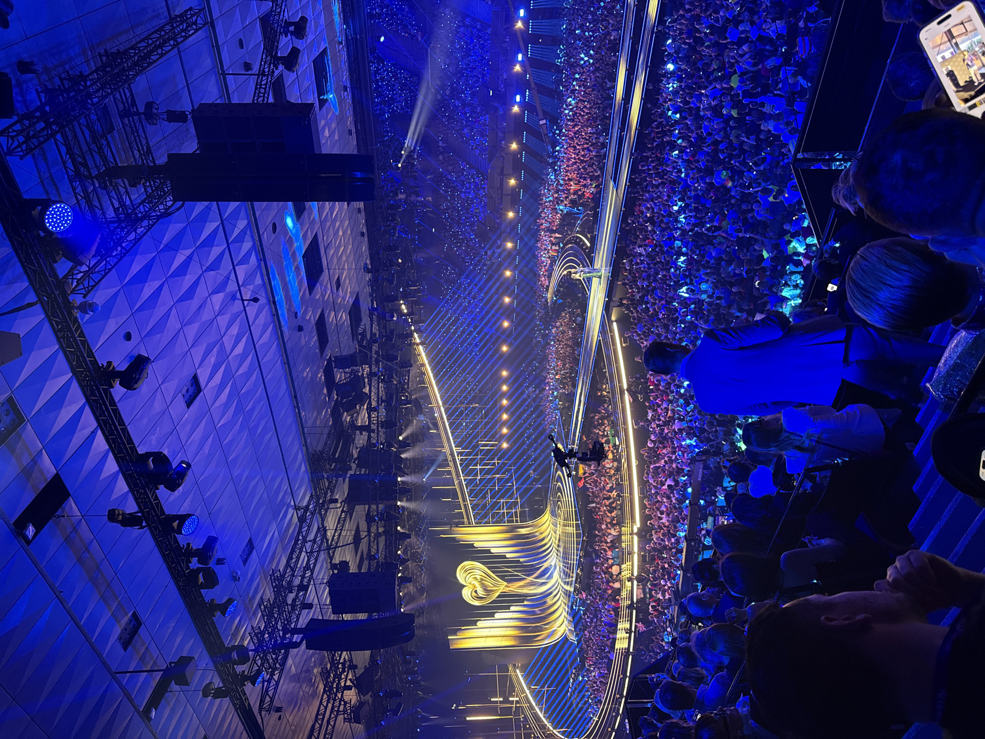 The image shows a large indoor venue filled with a dense crowd of people. The scene is lit with blue and golden lights. A stage with an elaborate golden structure that includes a heart design is visible. The overhead lighting and rigging are extensive. People in the foreground appear to be spectators watching the event. The photo was taken from an elevated perspective. The event takes place in Vienna, Austria.