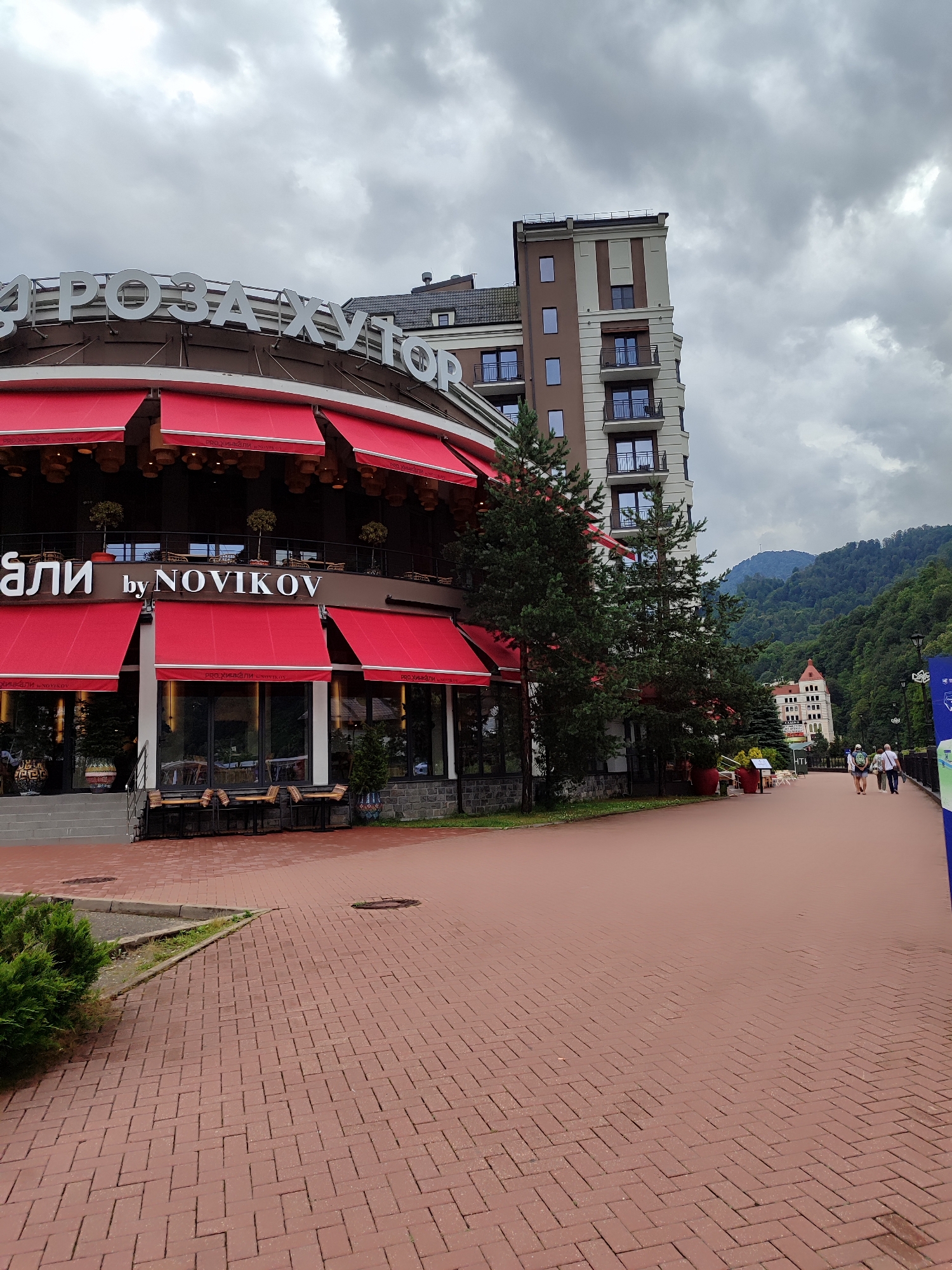This is a medium-shot, eye-level, vertical, outdoor, daytime photograph taken in Estosadok, Russia. The image shows a brick-paved walkway that curves to the right, leading away from the viewer. On the left is a circular building with a dark brown facade and red awnings extending over large windows. Above the windows, large white letters spell out "РОЗА ХУТОР" and "ХАЧАПУРИ by NOVIKOV". The building is partially obscured by evergreen trees. To the right of this building, a tall, modern, multi-story hotel with a beige and brown facade stands against a cloudy sky. In the background, a lush green mountainside rises, dotted with trees. Further in the distance, another building with a red roof is visible nestled amongst the trees. Several people are walking on the pathway in the middle distance, and a blue banner is visible on the far right edge of the frame. The overall atmosphere is overcast.