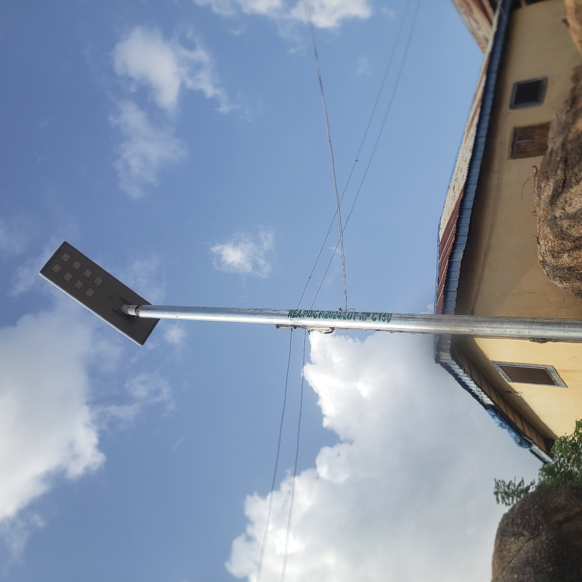 In Gwong, Nigeria, a solar-powered street light stands tall against a blue sky dotted with white clouds. The light, with its rectangular panel of LEDs, is mounted on a silver pole that bears green lettering reading "REAR/DIGITALIZATION/VOT/N150". Below the light, a yellow building with a corrugated metal roof, showing signs of rust and age, is visible. The building has several windows, some boarded up and others with dark panes. In the foreground, large, weathered rocks are scattered, and some greenery can be seen near the base of the building. Power lines are strung across the scene.