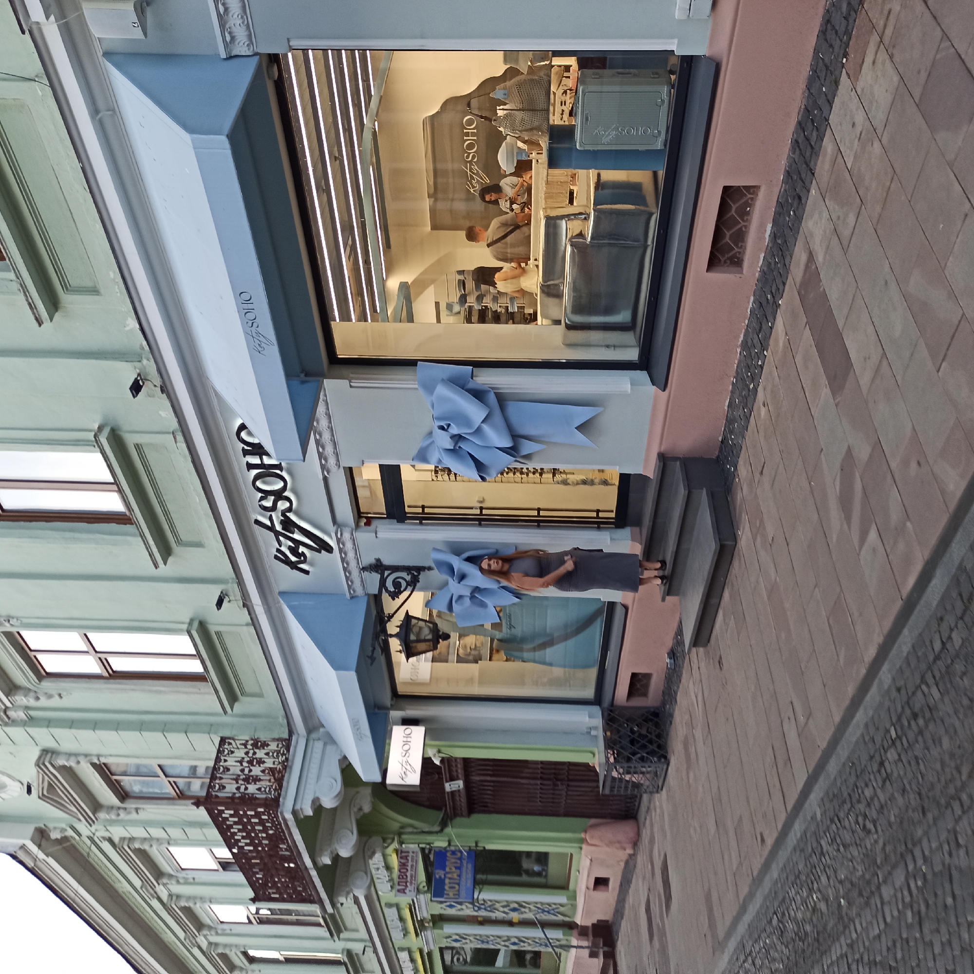 The image shows the exterior of a shop in Chernivtsi, Ukraine, identified by the signage "Katy Soho" visible on the storefront and an awning. A woman in a grey sleeveless dress and black heels stands on the steps leading up to the shop entrance, looking towards the camera. The shop's facade is light blue-green with ornate architectural details. Large light blue bows are attached to the wall on either side of the entrance. The shop window displays merchandise and customers inside. To the left of the "Katy Soho" shop, there are other storefronts, including one with Cyrillic signage that appears to be for a lawyer ("АДВОКАТ") and another with "НОТАРІУС" (Notary). The street is paved with cobblestones and large rectangular stones. A metal grate is visible on the sidewalk near the building's foundation.