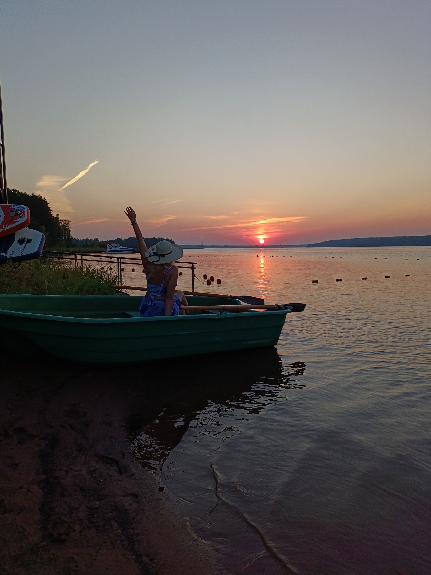 A woman in a wide-brimmed hat and a blue and white floral dress sits in a green rowboat on a body of water. She is facing away from the camera with her right arm raised, waving. The boat is near the shore, with a sandy bank visible to the left. In the background, the sun is setting over the water, casting an orange glow in the sky and reflecting on the water's surface. A line of buoys stretches across the water. Trees line the distant shore, and a small white boat is visible further out. Several colorful boards or kayaks are stacked on a rack near the shore to the left of the rowboat. The image appears to have been taken in the city of Chaykovsky, Russia, along the Kama River during sunset.