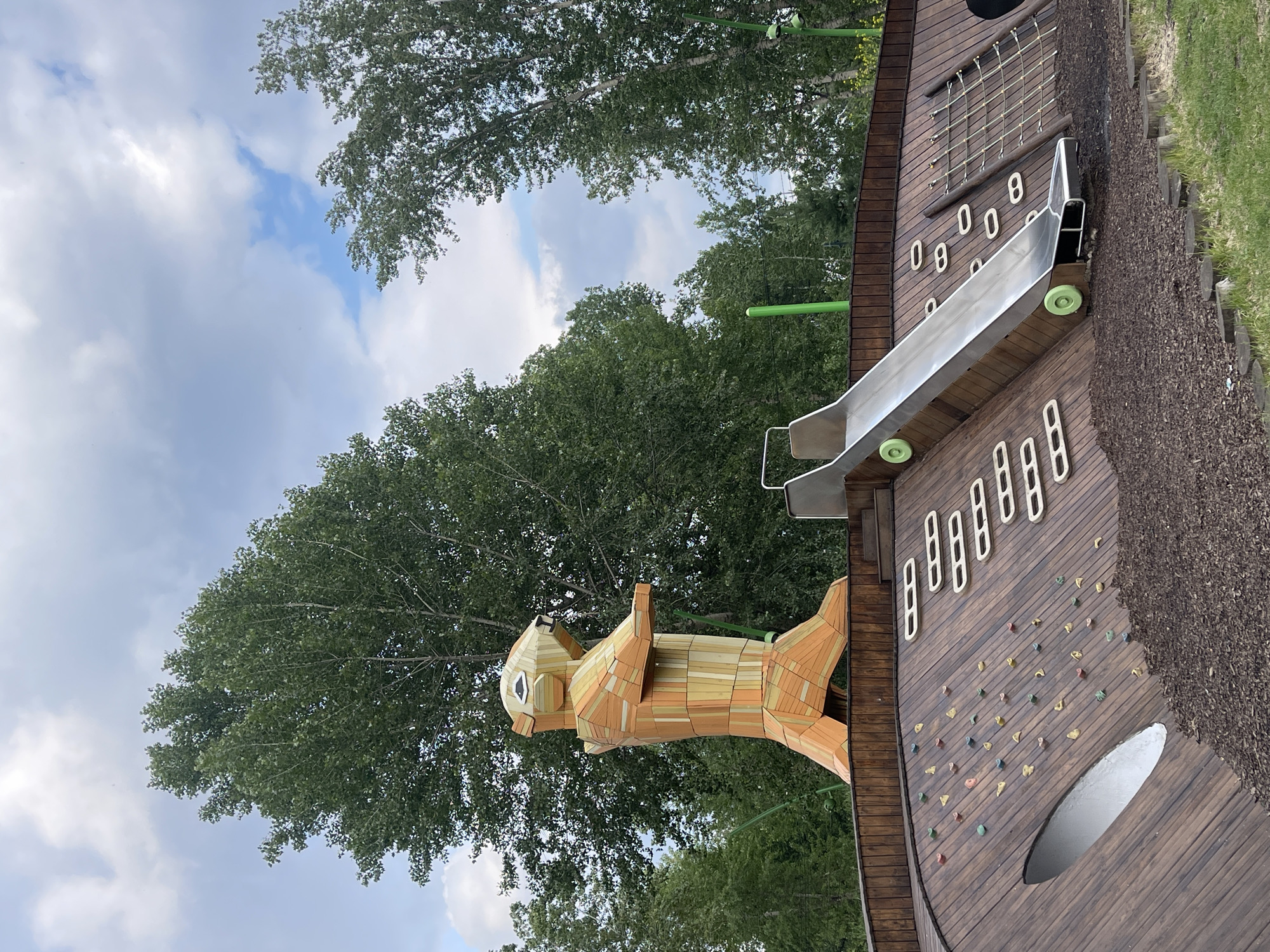 The image shows a wooden sculpture of a marmot in a park. The marmot is orange and yellow and is standing on its hind legs. It is holding its front paws out in front of it. The sculpture is part of a playground, which includes a slide, a climbing wall, and a rope net. The playground is made of wood and is surrounded by trees. The sky is blue with some clouds. The location is in Krasnoyarsk, Russia, at the "Park of the Gods" in the Grado-Mikhailovskaya embankment.