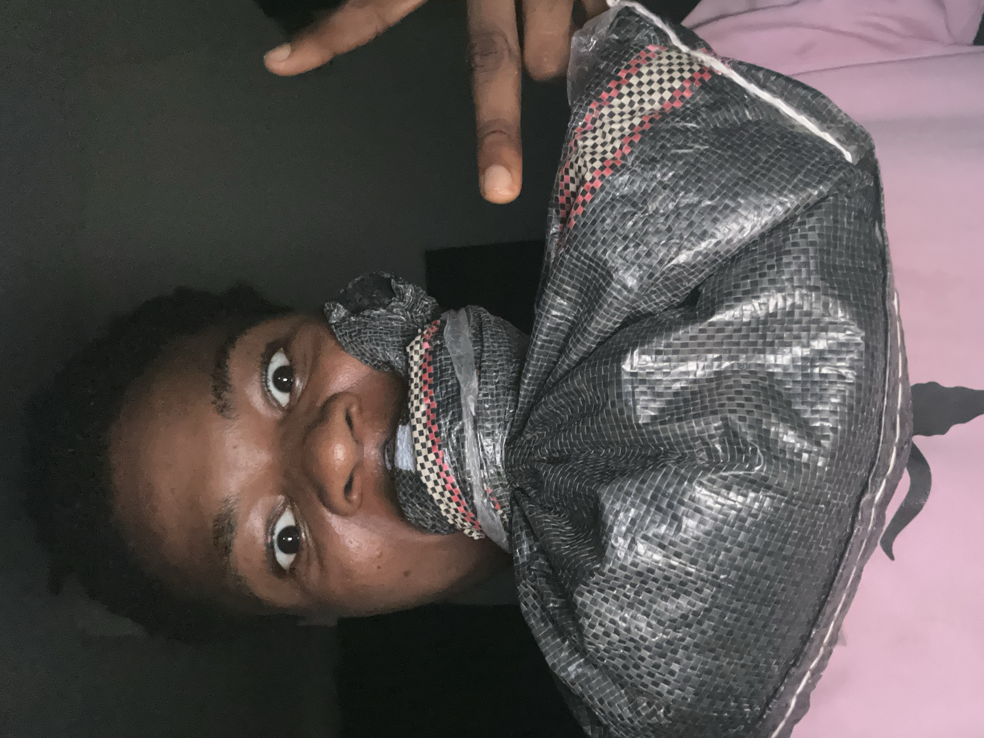 A person with dark skin and dark hair is shown from the chest up, looking directly at the camera with a surprised expression. Their mouth is covered by a black bag that is tied shut. The person is wearing a pink shirt. A finger is raised in the air to the right of their head. The background is dark and indistinct, with no discernible features to indicate a specific location within Ihiagwa, Nigeria.