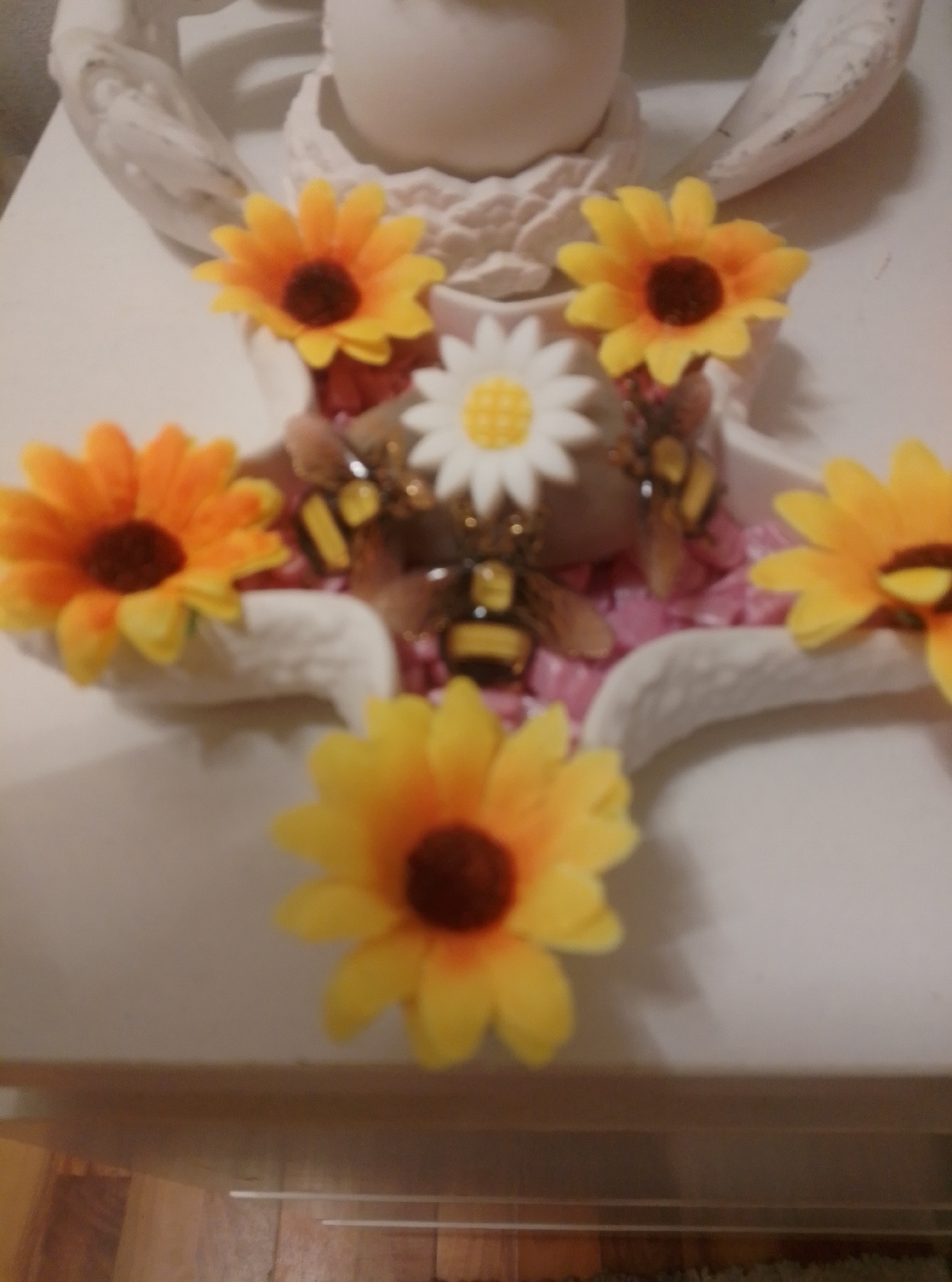 This is a close-up, eye-level, indoor photograph of a decorative arrangement. The arrangement features several artificial sunflowers, varying in size and color, with yellow petals and dark brown centers. Interspersed among the flowers are three decorative bee figurines, detailed with black and yellow stripes and translucent wings. The bees appear to be positioned on or around the flowers. The arrangement is set against a white surface, possibly a table or shelf, and partially rests on a white, curved base that resembles folded material or petals. In the background, out of focus, is a white, sculpted object with prominent wing-like protrusions, possibly part of a larger sculpture or decorative piece. The lighting is soft, casting subtle shadows. There is no indication of the location being in Wedel, Germany; the image provides no geographic context.