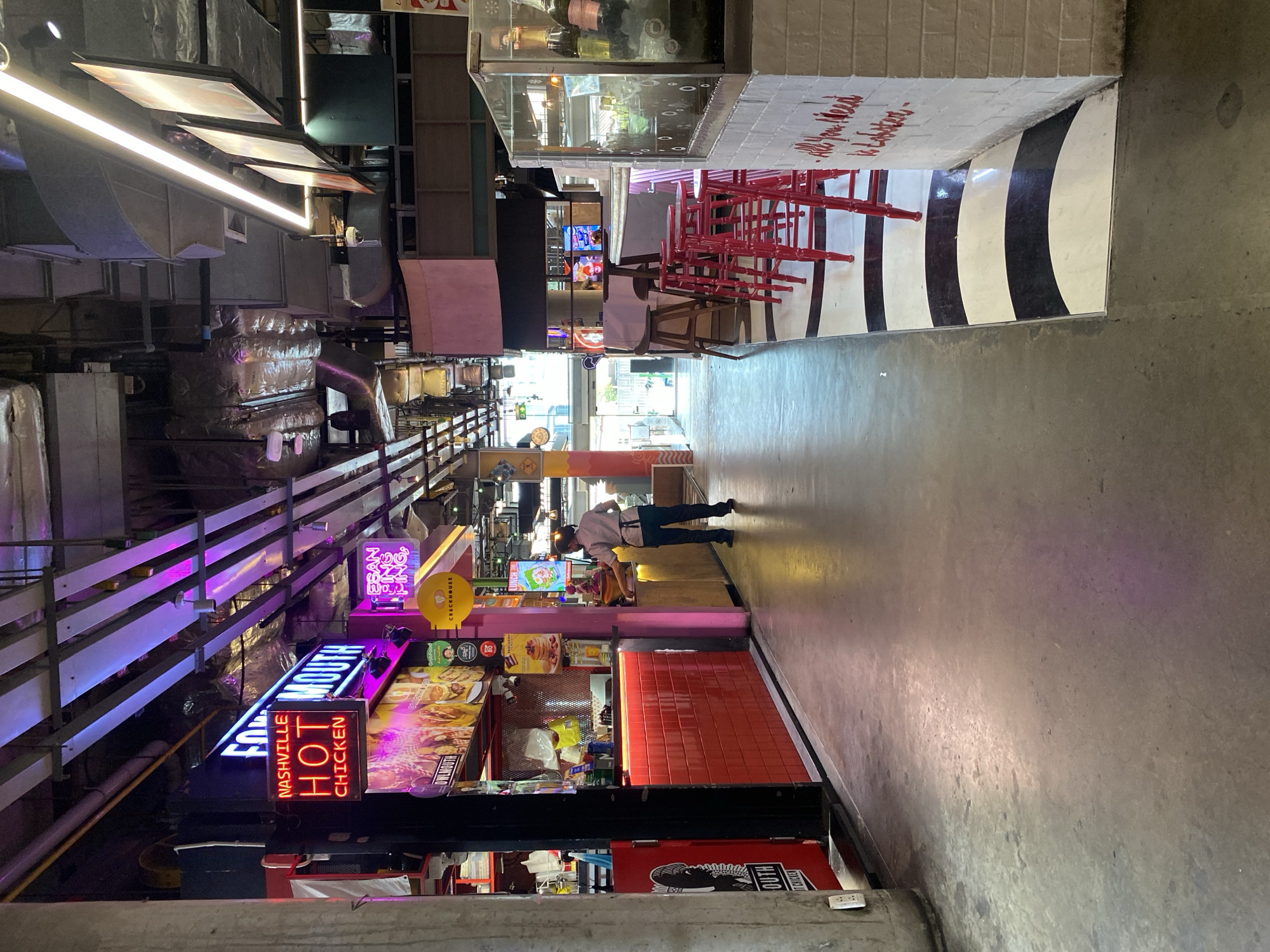 The image depicts a commercial hallway or passage in Bangkok, Thailand. The space is characterized by its concrete floor and a variety of food vendors lining the sides. A person is seen walking along the hallway, seemingly browsing the food options. The vendors are marked by brightly lit neon signs and displays of food items. The environment has a somewhat industrial aesthetic, with exposed structural elements and utilities visible overhead. The color palette is dominated by the neon signs, ranging from blue and pink to yellow and red.