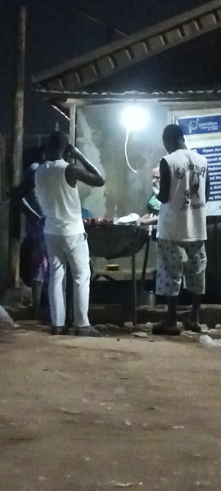 The image depicts an outdoor scene at night, possibly in Sabon Tasha, Nigeria. Three people are visible: two men are facing a grill, while another person is standing to the side. One man is wearing a white tank top and light-colored pants and is holding something to his ear, possibly a phone. The other man is wearing a white tank top with a design on the back and patterned shorts. A light is positioned above the grill. A small shed-like structure is in the background with a sign on the right side. The ground appears to be dirt or gravel.
