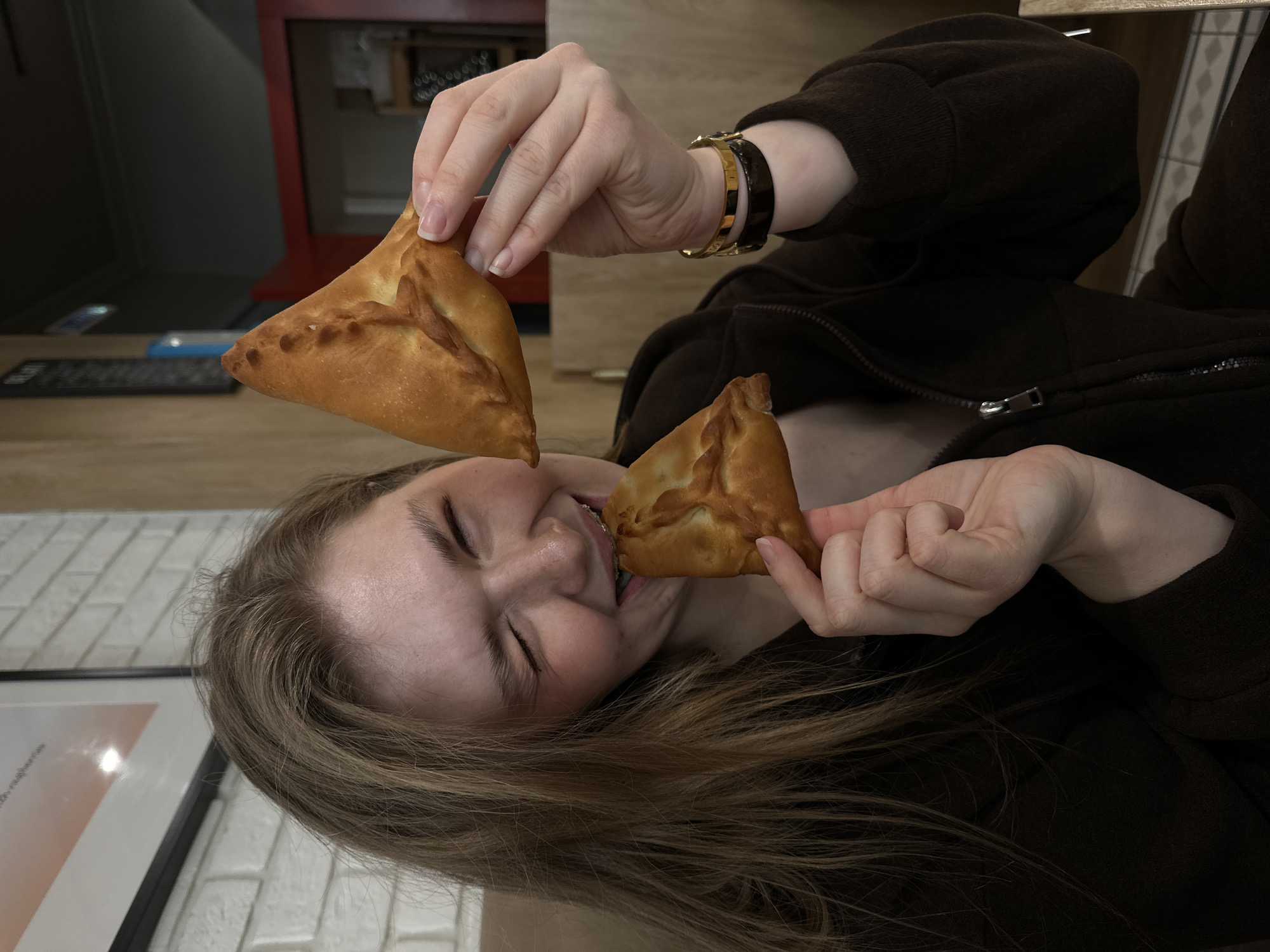 An indoor scene in what appears to be a Tatar cafe or restaurant in Kazan, Russia, features a young adult female. She has long, light brown hair and is wearing a dark brown zippered hoodie. On her left wrist, she wears two bracelets: one gold-toned, one dark brown. She is holding two golden-brown, triangular-shaped pastries, resembling *echpochmak*. She holds one pastry in her right hand and is biting into the second pastry held in her left hand. Her mouth is open, revealing upper dental braces, and her eyes are squinted in an expression of enjoyment or playful action. The background includes a white brick-patterned wall with a partially visible framed poster containing the Cyrillic word "малина" (raspberry), wooden paneling, and a blurred dark red doorway to the right.