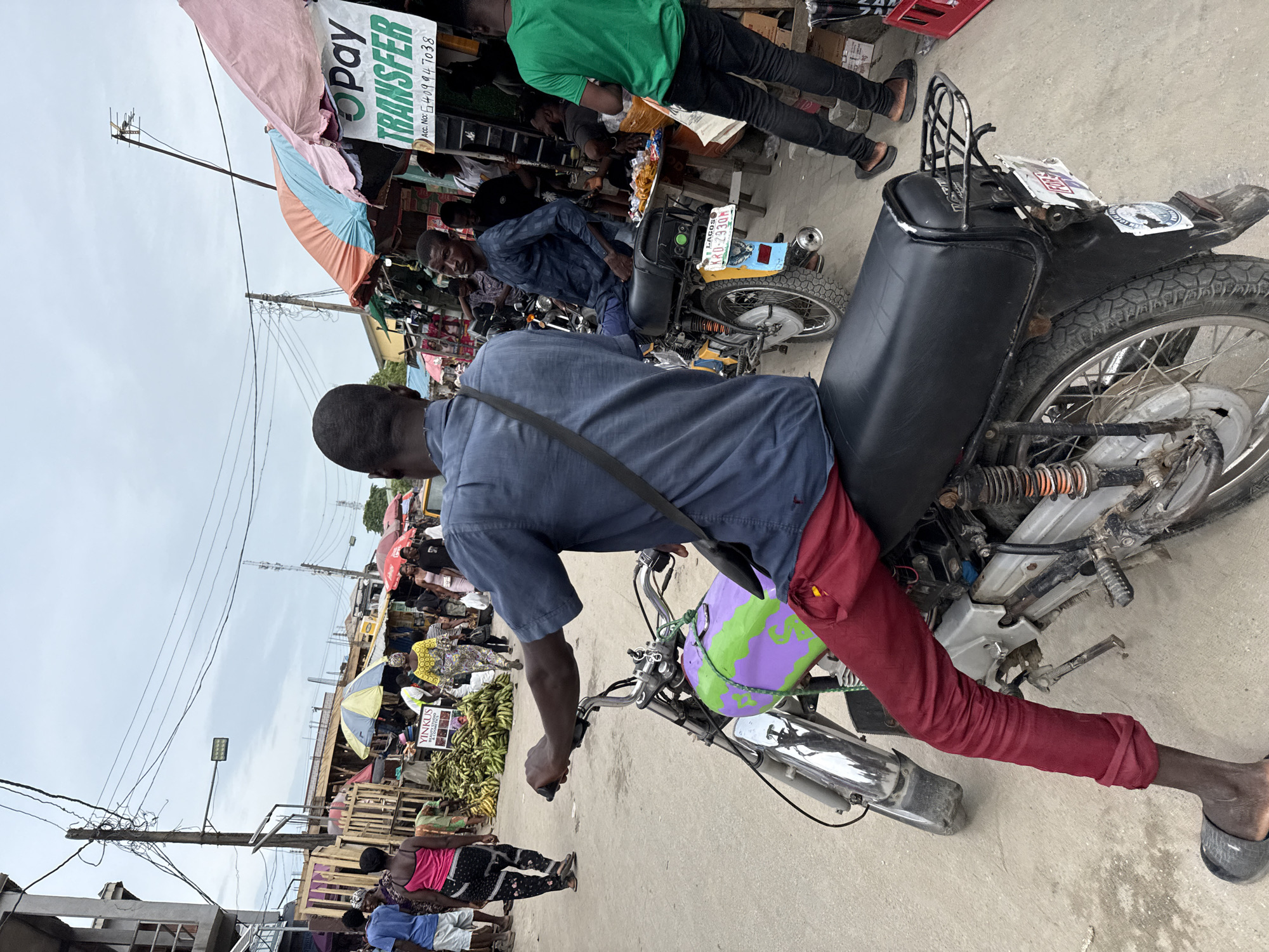 The image shows a man sitting on a motorcycle in what appears to be a bustling market area in Lagos, Nigeria. The man is wearing a blue shirt and red trousers. The motorcycle has a custom paint job of purple and green on the front. Behind him, there's a crowded street with vendors and shoppers. The storefronts have signs, one of which reads "OPay TRANSFER" with an account number. Other motorcycles are parked nearby, and vendors are selling various goods. The sky is overcast.