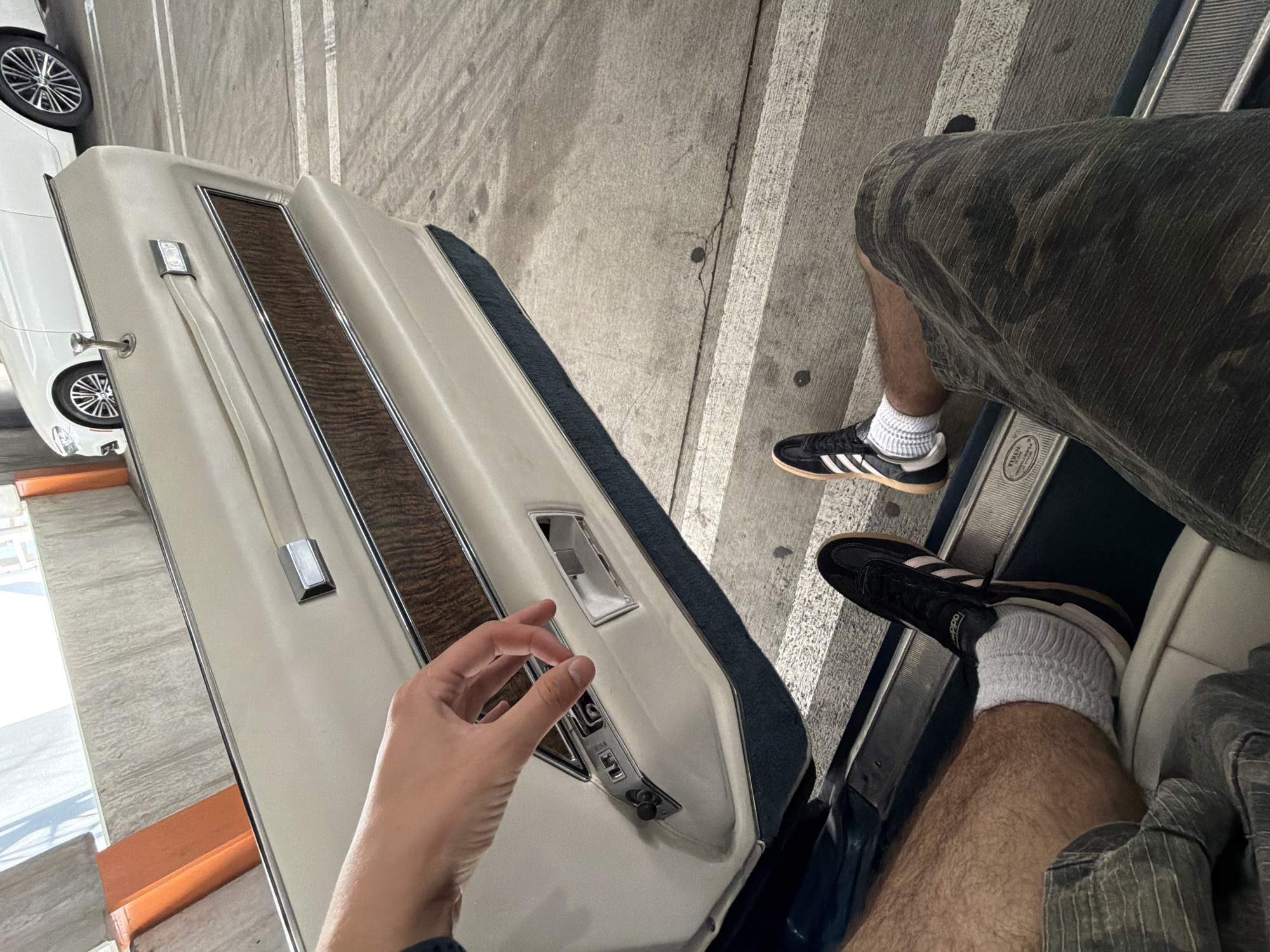 A first-person perspective shot captures a person's hands and legs from a seated position inside a car, with the car door open. The car interior features a cream-colored door panel with a wood grain accent and chrome trim. The person's left hand is visible, reaching towards the car's interior door handle mechanism. The person is wearing camouflage-patterned shorts, white socks, and black Adidas sneakers. The exterior view shows a concrete parking lot with white parking lines and a white car parked nearby. The overall lighting suggests an outdoor, daytime scene, possibly in a parking garage or an open-air lot, which could be located in Los Angeles, California, United States of America.