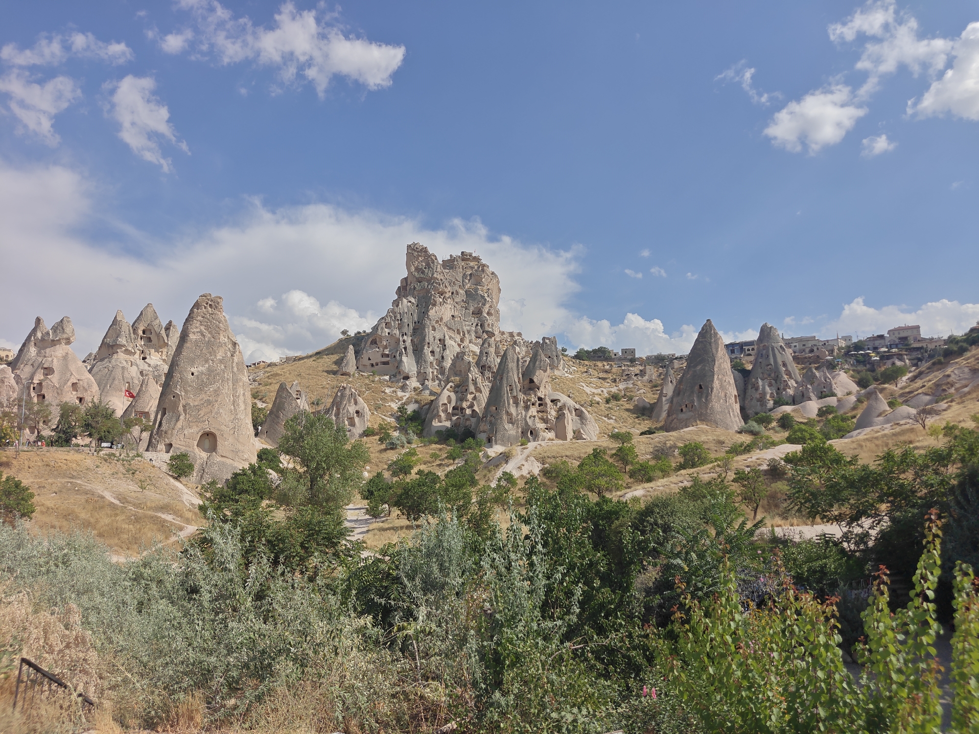 The image depicts the unique landscape of Uçhisar Beldesi, Turkey, characterized by a dramatic collection of fairy chimneys and rock-cut dwellings. These formations, carved by erosion over millennia, dominate the middle ground and background. Many of the rock structures feature openings, indicative of past human habitation or current usage as storage or shelters. In the foreground, lush green foliage and dry, sparse vegetation create a contrasting texture to the rocky terrain. The sky above is a vibrant blue, dotted with scattered white clouds, suggesting fair weather. In the distance, a few buildings are visible on the hillsides, integrated into the natural contours of the land. The overall scene conveys a sense of ancient history and the striking geological features of the region.