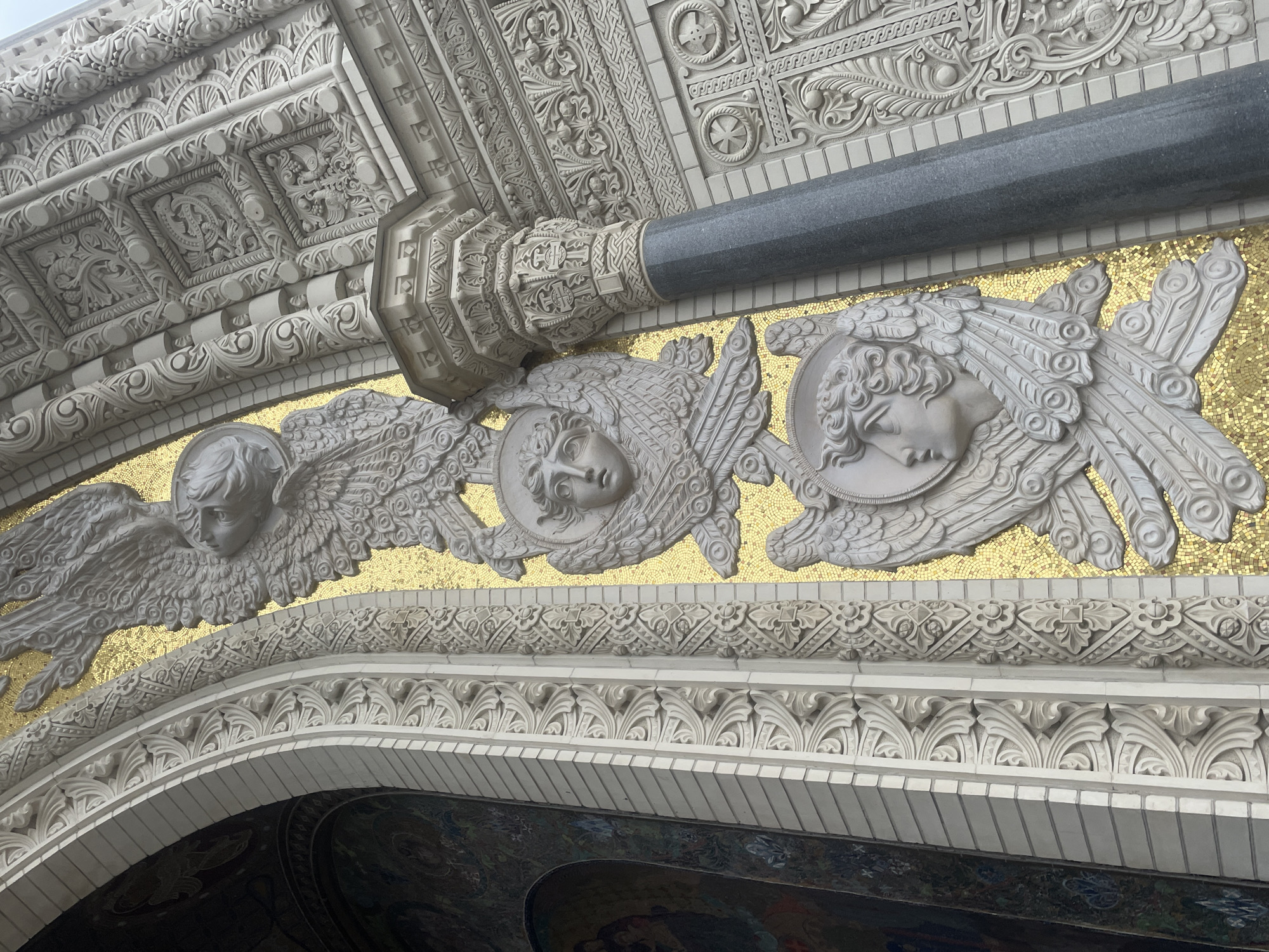 The image shows a close-up view of the exterior architecture of the Naval Cathedral of Saint Nicholas in Kronstadt, Russia. The focal point is a section of the building's facade featuring intricate relief sculptures of angelic figures against a gold mosaic background. One prominent sculpture depicts a winged angel's head and upper body, with detailed feathers and a halo. Below this, another similar but smaller angelic head is visible. The sculptures are made of white stone and are highly ornate, with fine detailing in the hair, wings, and facial features.
Flanking the angelic sculptures are decorative architectural elements. To the left, a section of an arched entryway is visible, adorned with relief carvings and partially obscured by a mosaic mural. To the right, a substantial grey granite column stands, topped with a capital featuring elaborate carvings. The rest of the facade to the right of the column is covered in complex, multi-layered stone carvings, including repeating patterns, square panels with smaller reliefs, and circular motifs. The overall impression is one of grandeur and meticulous craftsmanship, characteristic of late 19th and early 20th-century religious architecture. The lighting suggests it is daytime, possibly overcast, as there are no strong shadows.