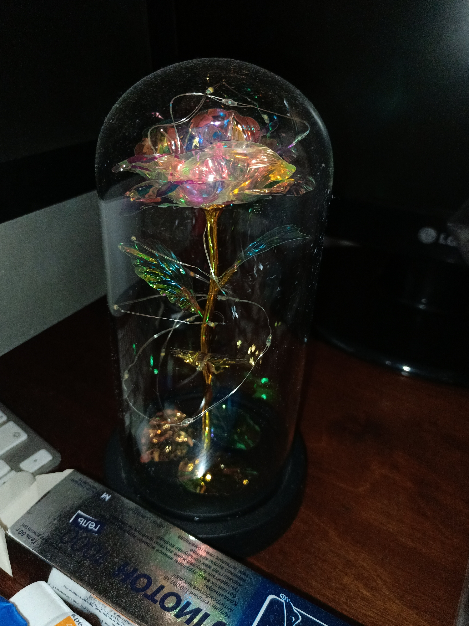 The image displays a decorative artificial rose encased in a glass dome, illuminated by tiny fairy lights. The rose appears to be made of iridescent material, reflecting various colors. The glass dome has a slight film on its surface, suggesting it may be dusty. The dome sits on a black circular base.
In the foreground, to the left, a portion of a product box with Russian text is visible. The text on the box includes "ИМОТОК 1000", "гель", and "M". Further left, a silver keyboard is partially visible. The background is dark, with a hint of a dark surface that might be a desk. A portion of an LG electronic device with its logo is also visible on the right side of the image. The lighting suggests an indoor setting, likely during the evening or in a dimly lit room, as the fairy lights are prominent. There are no people in the image. The overall scene evokes a sense of decorative display or personal adornment.