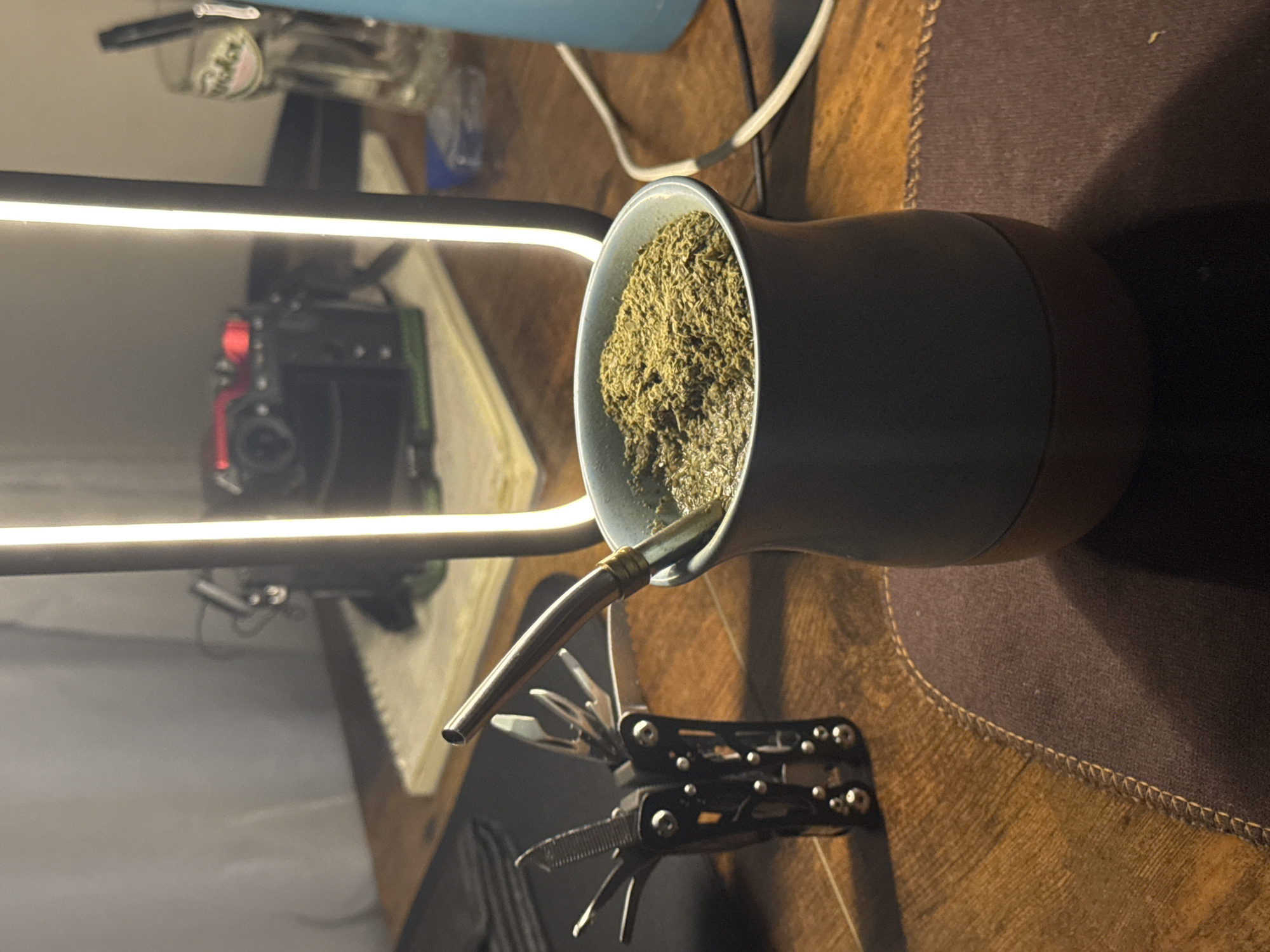This is a close-up, eye-level shot of a yerba mate gourd with a metal bombilla in a dimly lit indoor setting, likely a desk area. The gourd is filled with yerba mate and hot water. To the left of the gourd is a black multi-tool with various implements extended. In the background, a bright, vertical LED lamp illuminates a portion of the scene. Reflected in the lamp's surface are a camera and a glass with a pen inside. The objects are placed on a wooden desk with a textured brown mat beneath the gourd. There is no indication of the specific location in Lâm Viên - Đà Lạt, Vietnam, as the image lacks any identifying landmarks or context specific to that city.