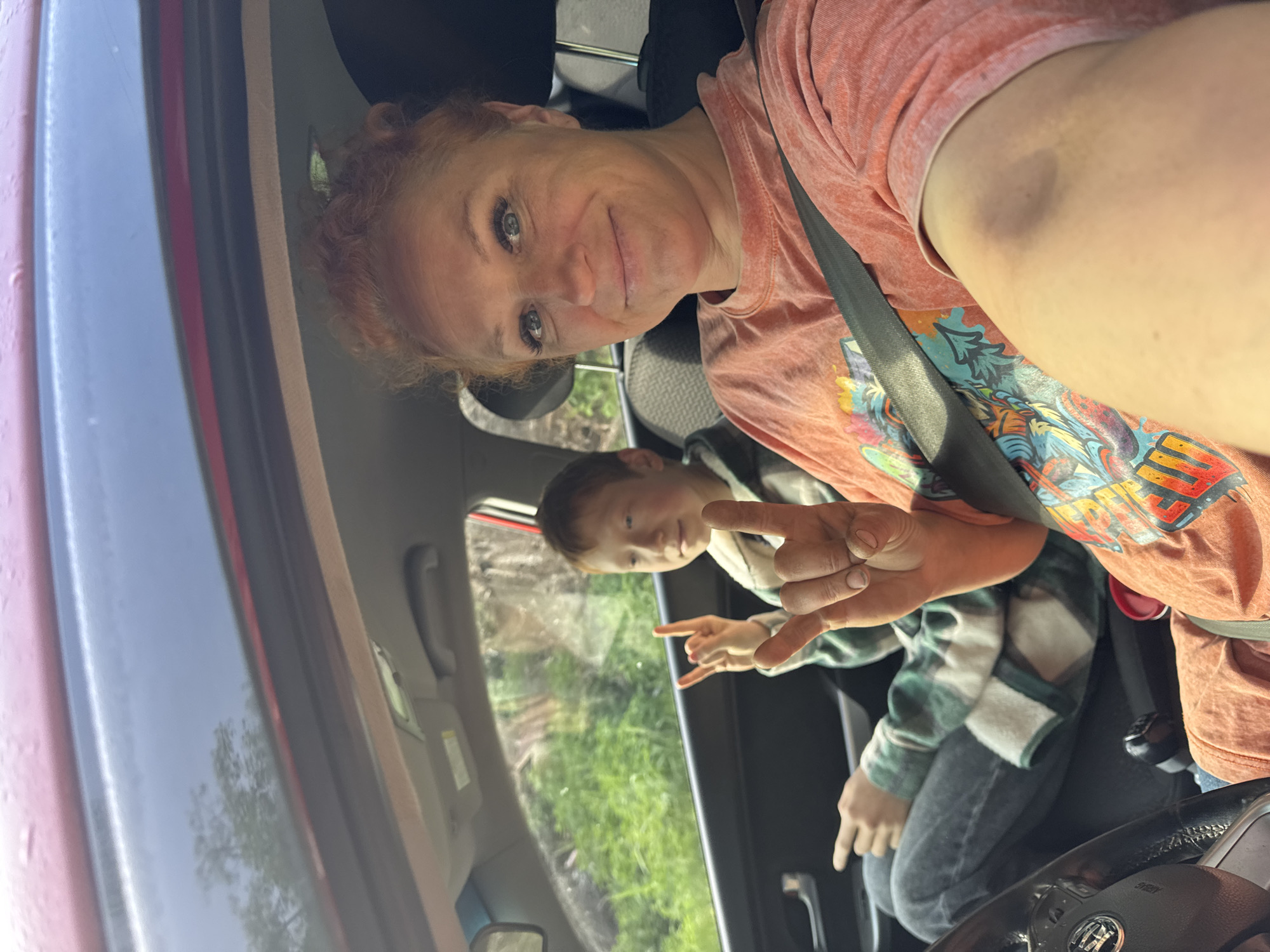 A selfie was taken from inside a red vehicle. A female with red hair and a peach-colored shirt is in the driver's seat wearing a seatbelt. The second person, a male child, is sitting in the back seat, wearing a green and white flannel shirt and jeans. Both subjects are facing toward the camera, throwing up what appears to be a "rock on" hand gesture. The child is positioned near the rear passenger window. The backdrop indicates a natural green scene. The location is not possible to determine.