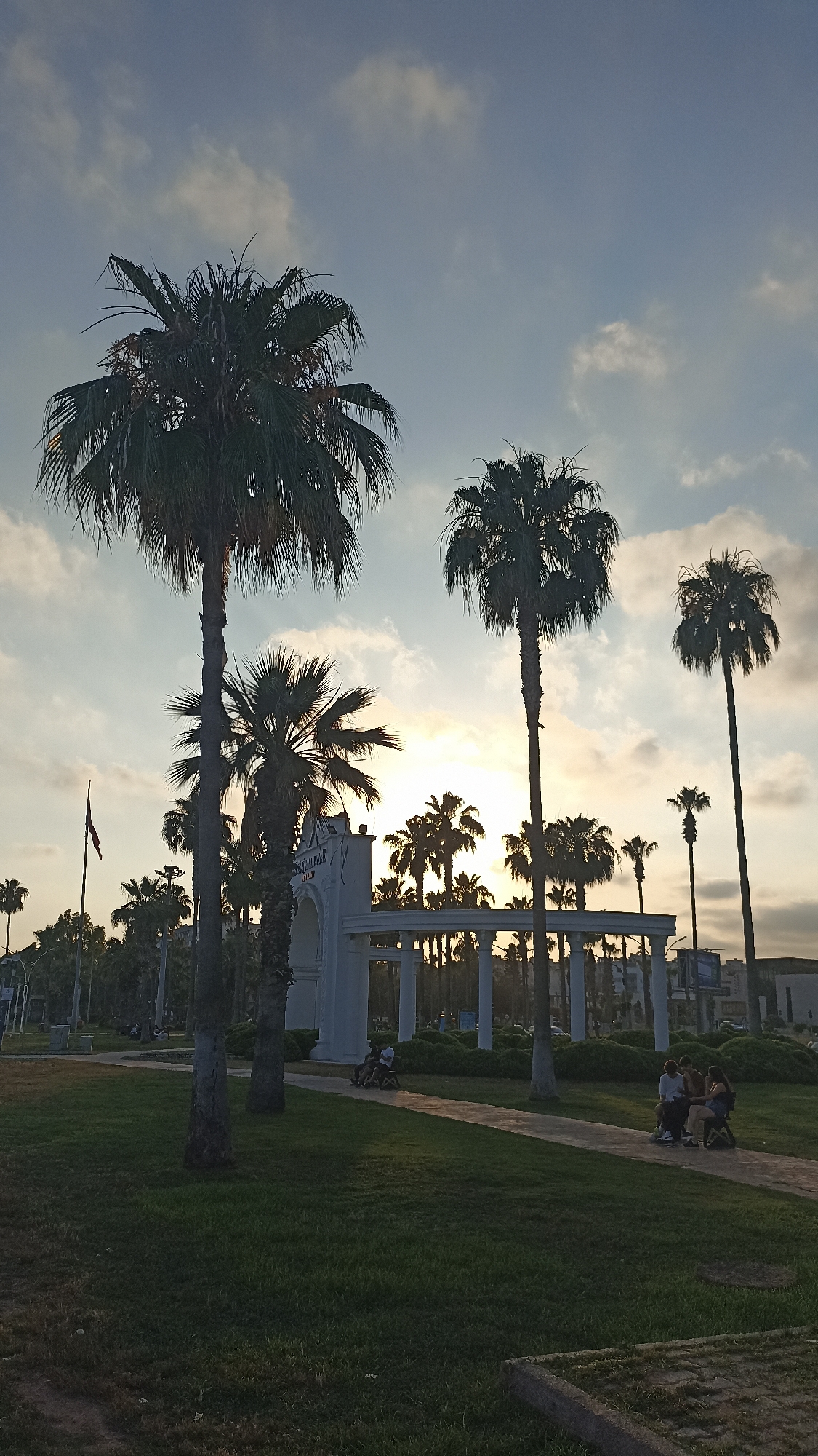 This image captures a sunset scene in Yenişehir, Turkey, featuring a park-like setting with numerous palm trees silhouetted against a softly clouded sky. The sun is low on the horizon, casting a warm glow that is visible through the trees and illuminating the clouds.
In the foreground, a lush green lawn leads to a paved pathway. Several palm trees of varying heights are scattered throughout the scene, with one particularly tall, slender palm tree standing prominently on the left side of the image.
Further into the park, a white architectural structure with columns and arches is visible. Behind this structure, more palm trees create a dense backdrop.
On the pathway, a small group of people are seated on what appear to be benches or low platforms. They are casually interacting, suggesting a relaxed atmosphere. One person is riding a small wheeled vehicle.
A flag pole with a flag is visible on the left side of the image, near the entrance to the park. The overall mood is peaceful and serene, typical of a late afternoon or early evening in a pleasant outdoor location. There is no discernible text visible in the image.