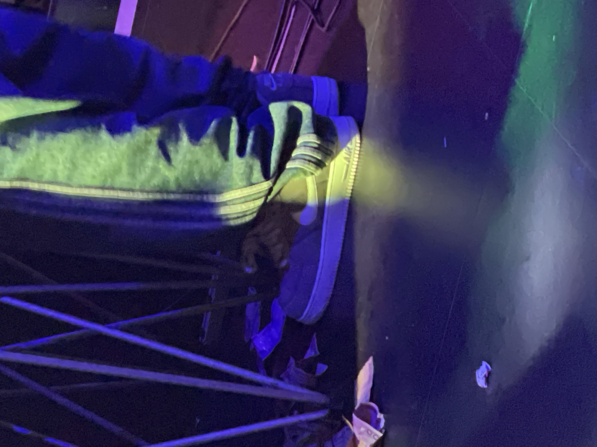 A person wearing gray Nike Air Force 1 sneakers and a light green and black jacket is standing behind a blue metal railing. Debris is scattered on the ground near the feet. No information about the location being in Obosi, Nigeria, is available in the image.