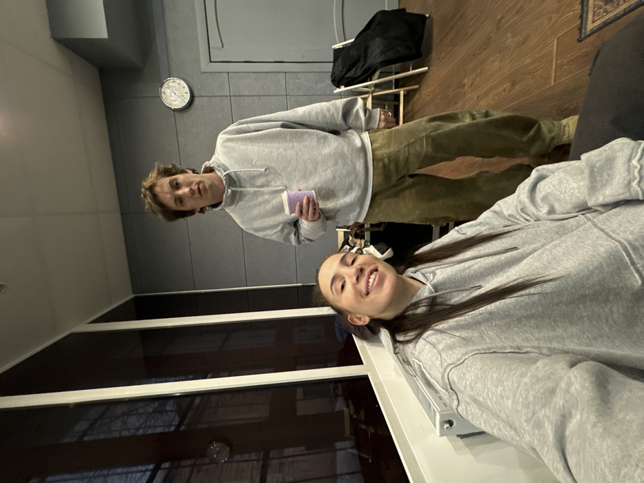 The scene is an interior room within an unidentified building in Moscow, Russia, resembling a studio or modern office. The left wall features large dark-framed windows reflecting exterior buildings and a dim sky. A white radiator is positioned below these windows. The back wall is grey, possibly paneled, with a light grey door on the right and a circular analog wall clock indicating approximately 1:55. The floor is light brown wood laminate, with a segment of a dark patterned rug visible in the bottom right.
Two individuals are present. In the foreground, a female with long dark hair, wearing a light grey hooded sweatshirt, is seated and smiling directly at the camera. Behind her, a male with wavy light brown hair stands, holding a light purple disposable cup in his right hand. He wears a light grey long-sleeved quarter-zip sweatshirt with a small dark logo and olive green wide-leg trousers, maintaining a neutral expression. In the background on the right, two folded chairs are visible, one with a black jacket draped over it. Indistinct dark objects are also visible behind the standing male.