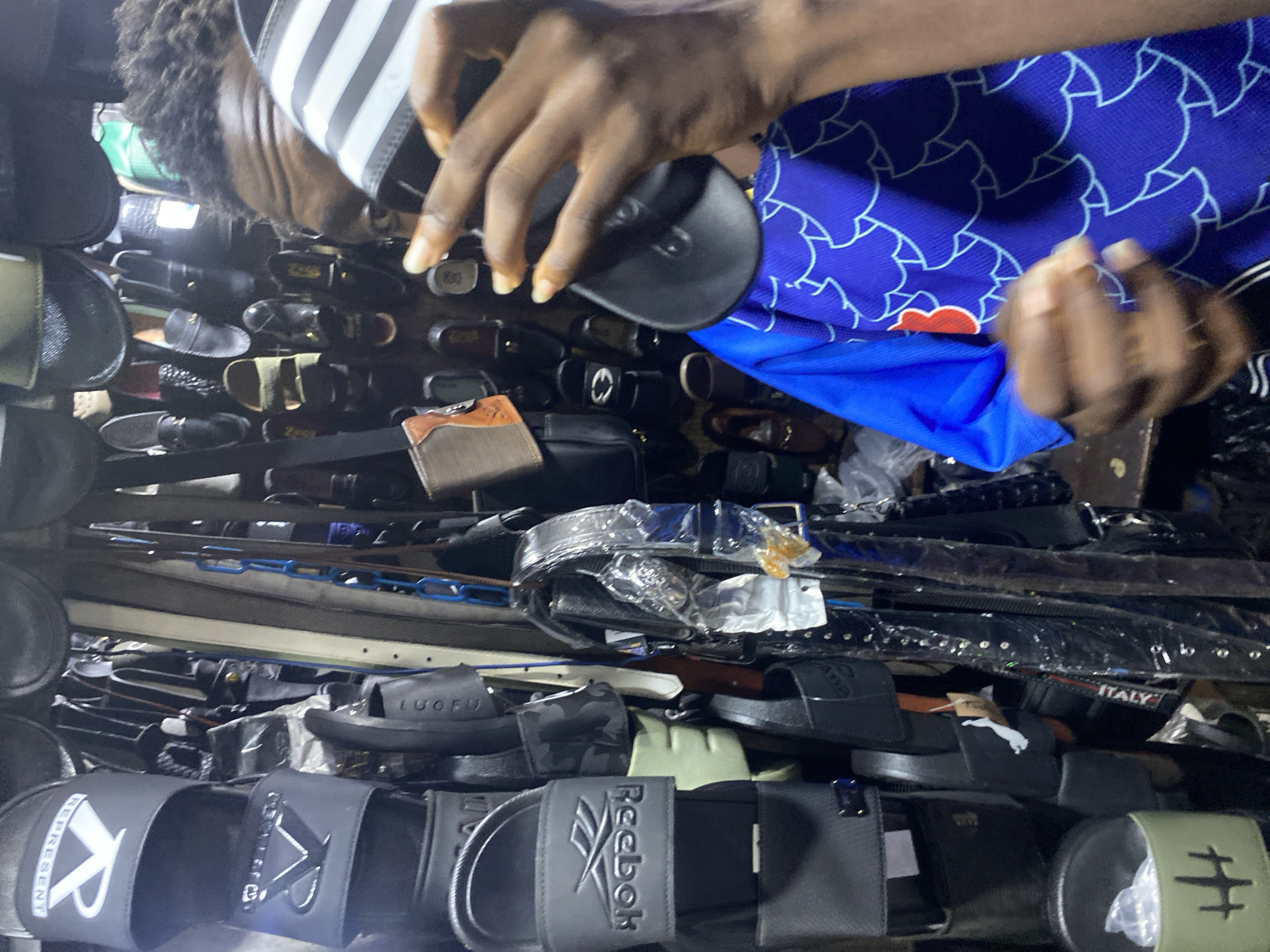 In Lagos, Nigeria, a person with dark skin and short, dark, curly hair is holding two sandals above a densely packed display of footwear and belts. The person is wearing a blue shirt with a patterned design. The sandal in the left hand has white stripes on a black background, while the other sandal is entirely black. Below, a variety of sandals are visible with different brands and designs, including Puma, Reebok, and a sandal with the inscription "ITALY." There are also multiple belts wrapped in plastic.