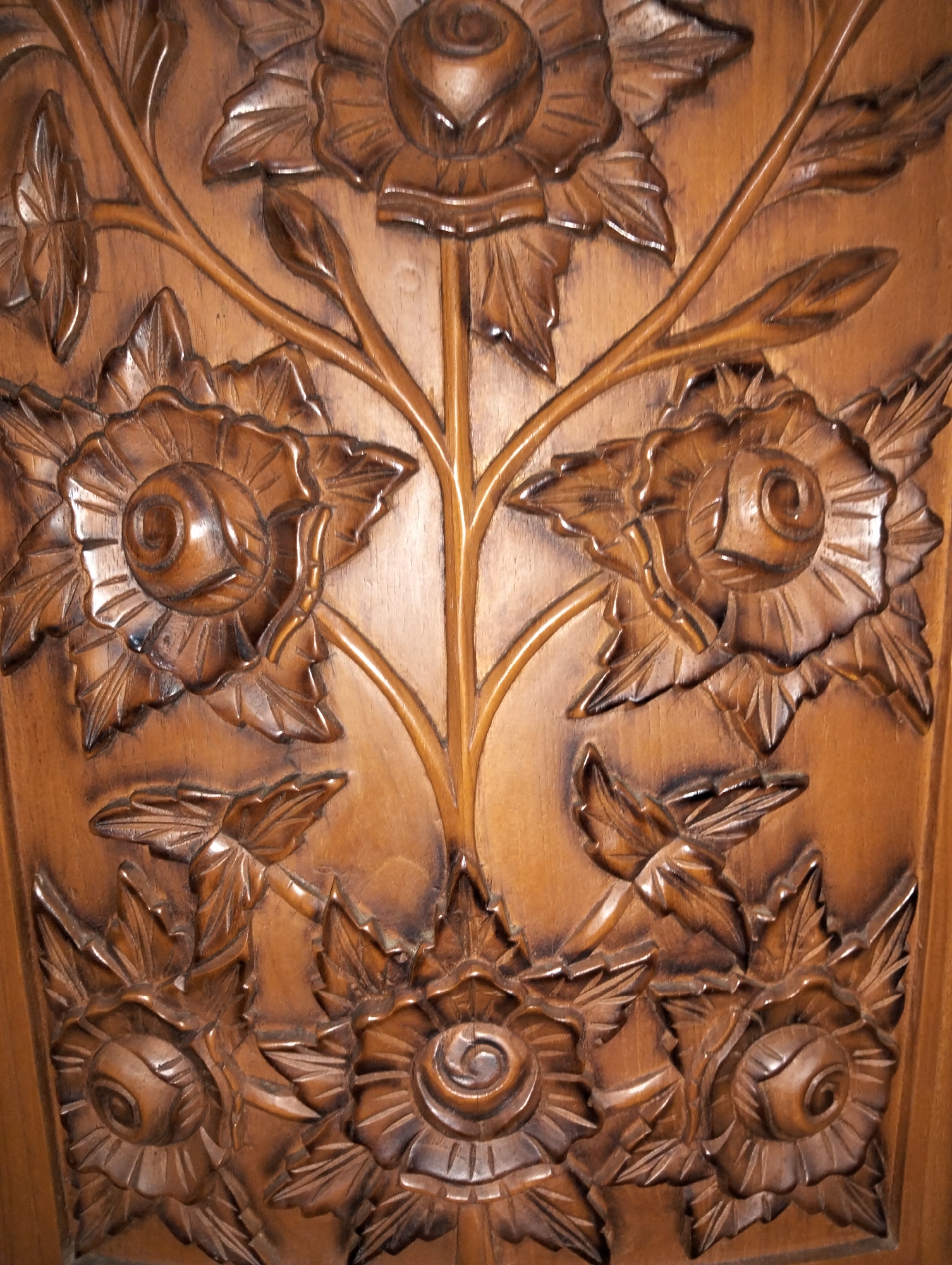 The image displays a detailed wood carving of floral motifs, featuring stylized roses and leaves with intricate petal and vein textures. The carving is rendered in a warm, medium-brown wood, exhibiting a polished finish that highlights the sculpted details and creates subtle shadows. The arrangement suggests a decorative panel or surface, with vines and floral elements interconnected in a flowing design. The artwork appears to be an ornamental piece, possibly part of furniture or architectural decoration. No specific location in Payagaboh, Indonesia, can be identified from the image alone, as it focuses solely on the carved object. There are no people, actions, or interactions depicted.