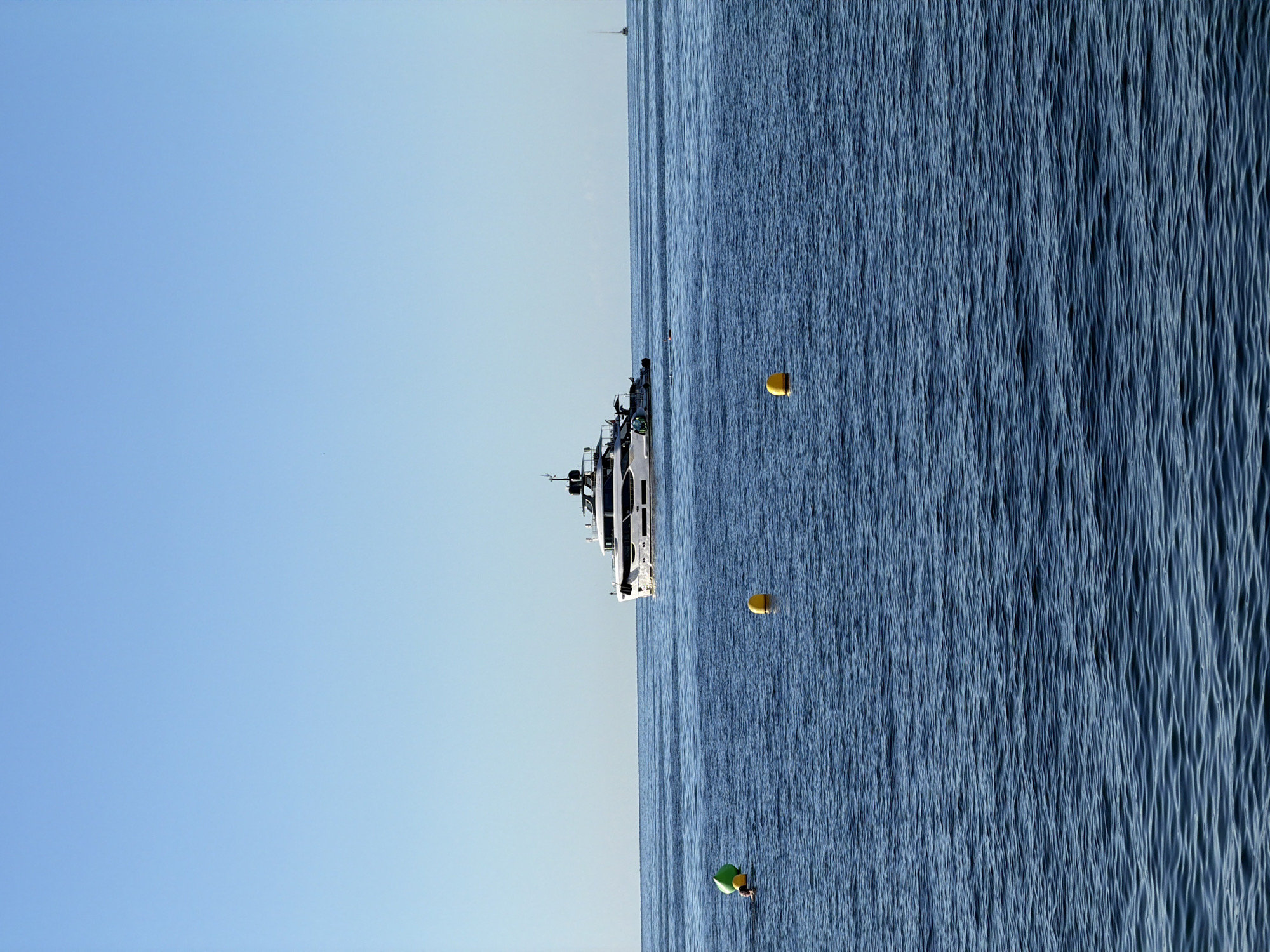 The image depicts a bright, clear day with a vast expanse of calm, deep blue ocean stretching to the horizon under a pale blue sky. In the mid-distance, a large, white luxury yacht with multiple decks and dark accents is the prominent subject, sailing from left to right. To the left of the yacht, in the foreground, a person is partially visible in the water, appearing to be swimming or treading water near a green and a yellow buoy. Further to the right, a single yellow buoy floats on the water's surface. In the far distance, a small, silhouetted sailboat is visible on the horizon. The overall impression is one of a serene, sunny day at sea, likely off the coast of Palmanova, Spain, given the context of the request.