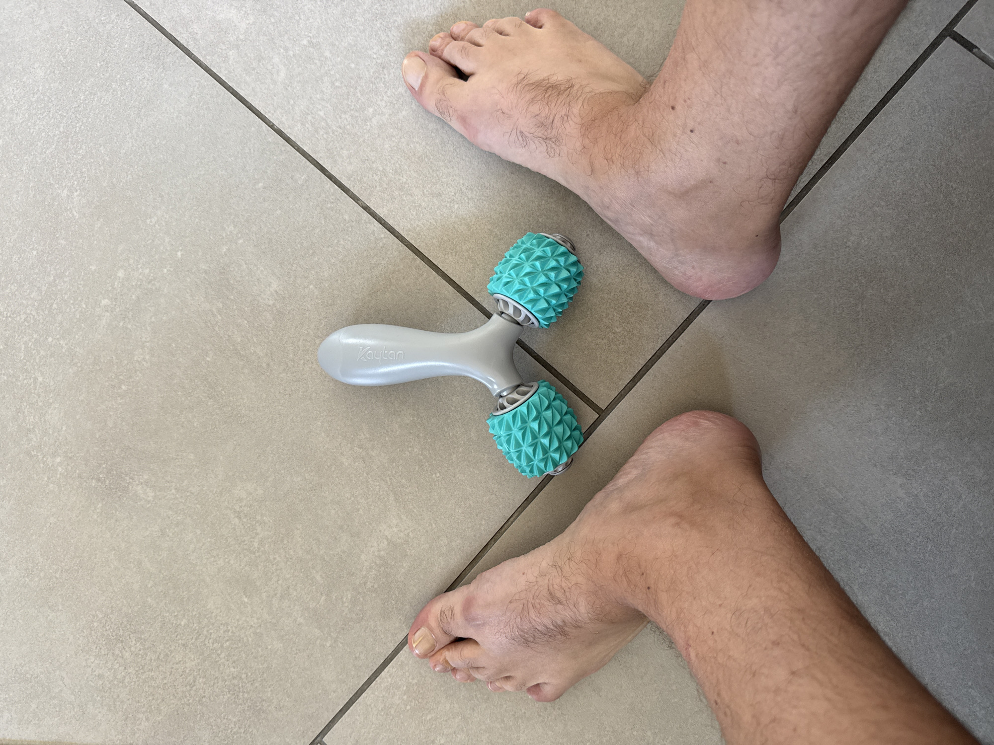 An overhead, wide shot captures a personal foot massager positioned between two bare feet on a light gray, tiled floor. The massager is a light gray handheld device with two turquoise, spiked rollers. The feet, one on the left and one on the right, are positioned facing upwards, with the toes slightly spread. The floor tiles are laid in a grid pattern, with dark gray grout lines. The lighting is bright and even, suggesting an indoor setting. No other individuals or objects are visible in the frame. The location of Drumettaz-Clarafond, France is irrelevant to the visual content of the image.